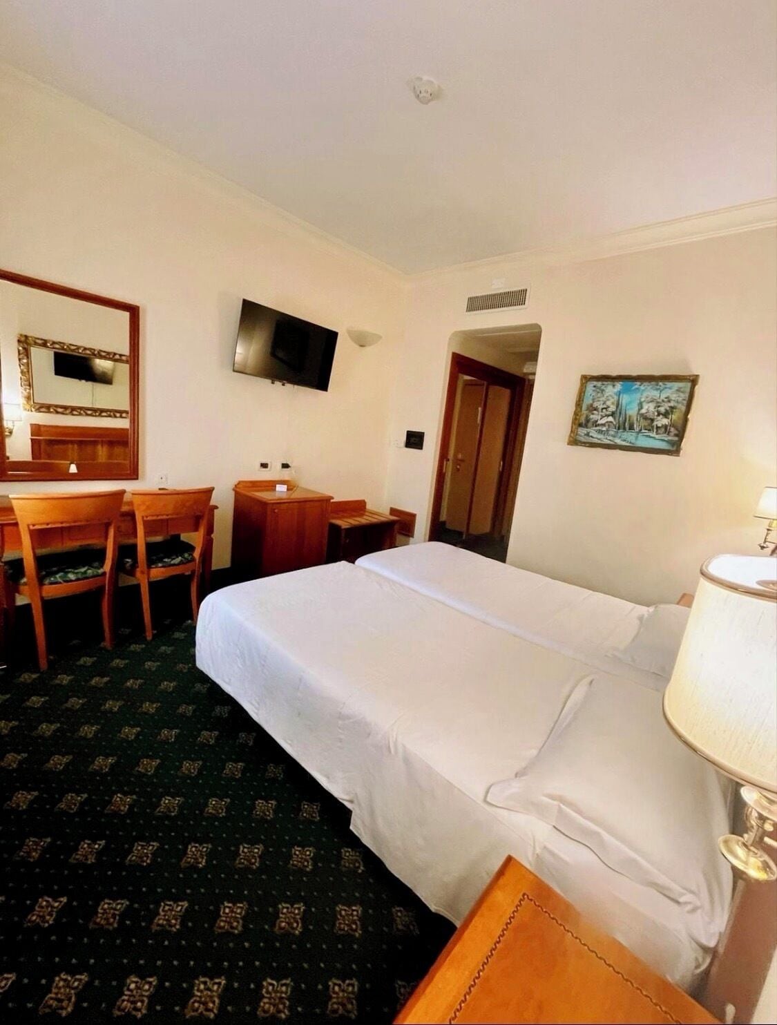 A hotel room with a bed, a desk with a chair, a television, and a painting on the wall.