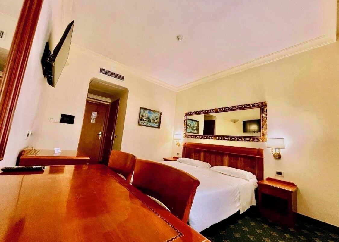 A hotel room with a wooden desk, a bed with white linens, a large mirror, and a nightstand with a lamp.