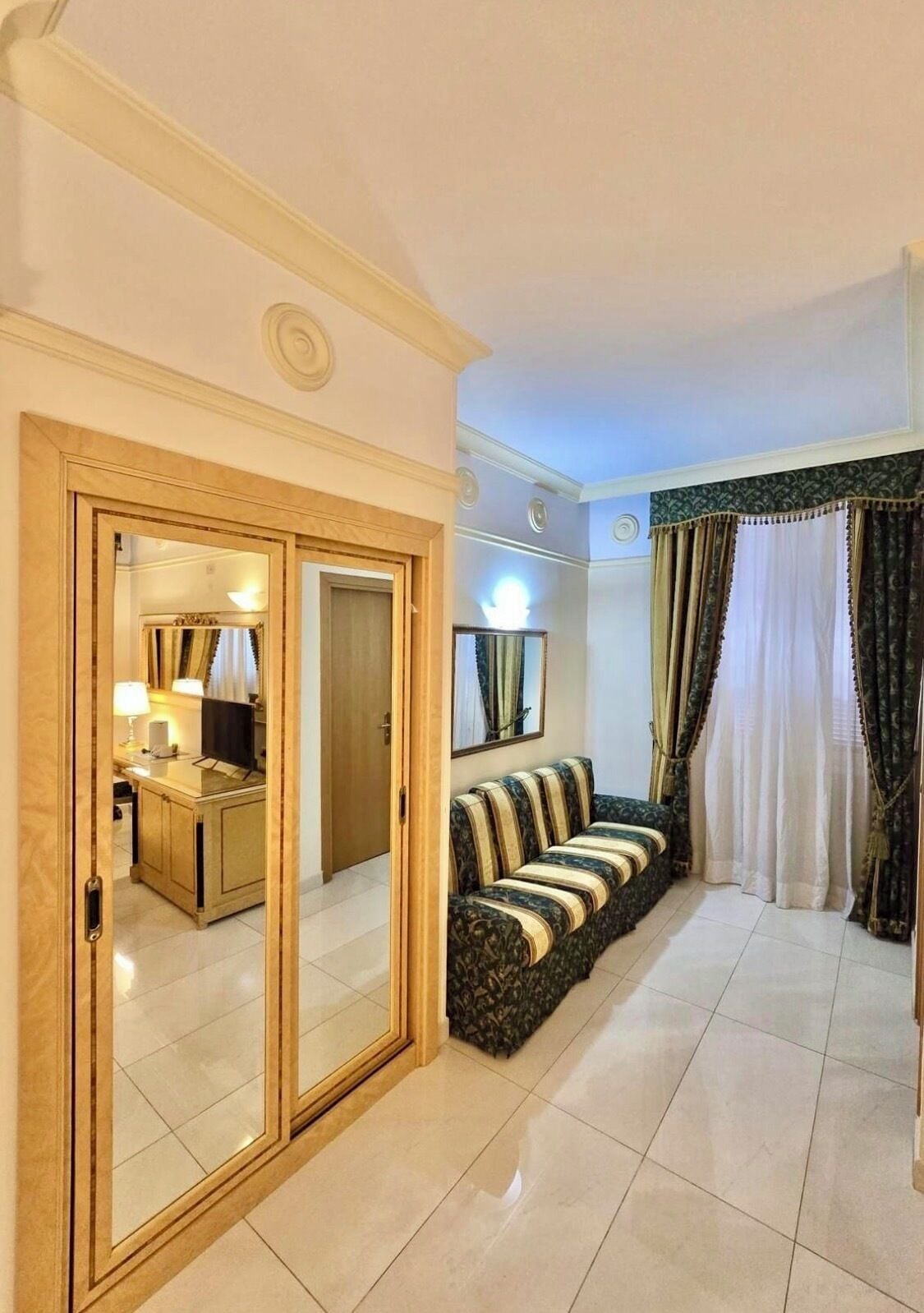 A hotel room with a mirrored wardrobe, a sofa, a television, and a dining area with a lamp.