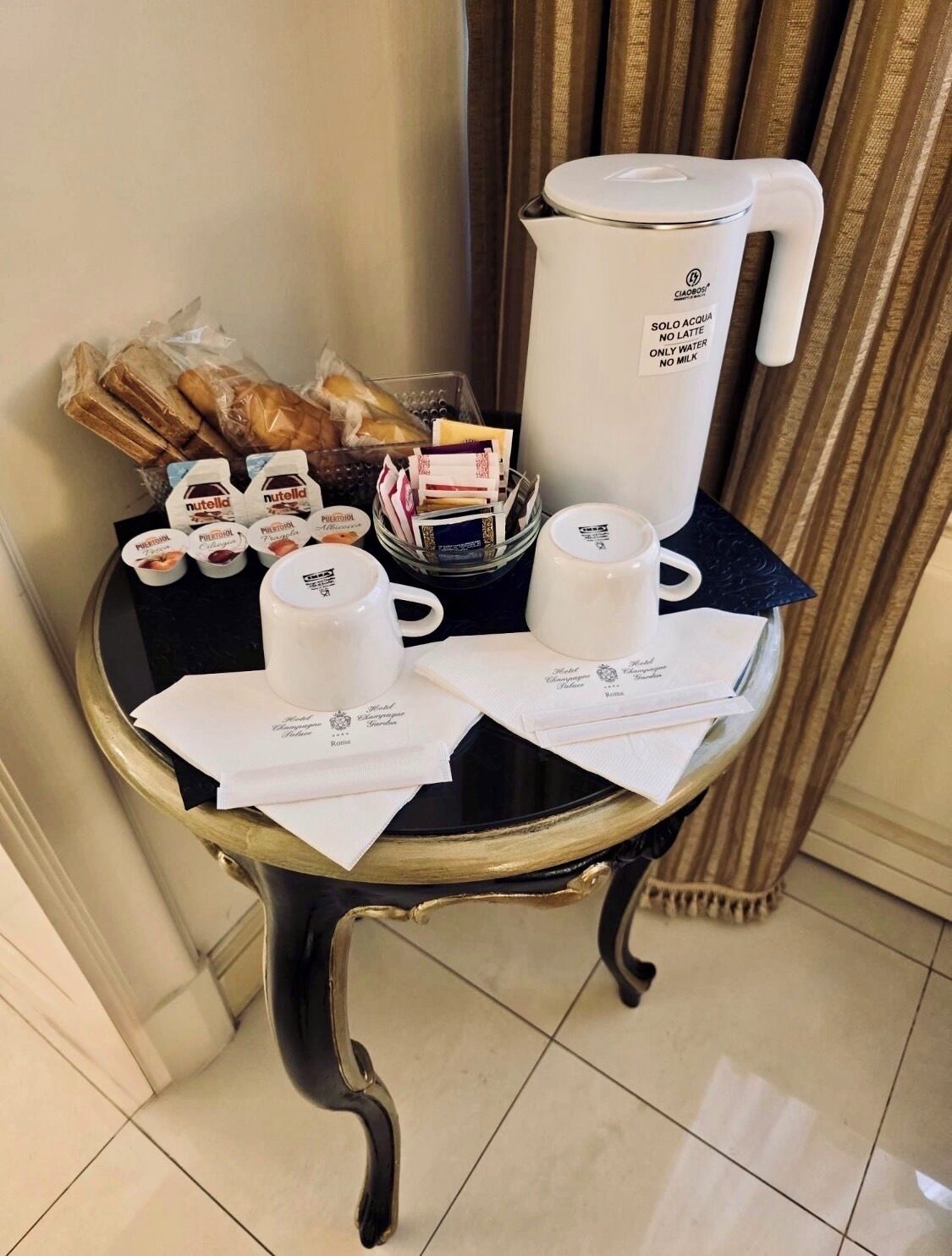 A table with a coffee maker, cups, Nutella jars, and pastries.