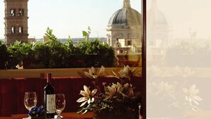 A table with a bottle of wine, a glass, and a vase of flowers, overlooking a cityscape with a tower and a dome. - Champagne Palace Hotel (Rome)