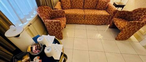 Un salón con un sofá estampado, una mesita auxiliar con una lámpara y una bandeja con comida y papeles.