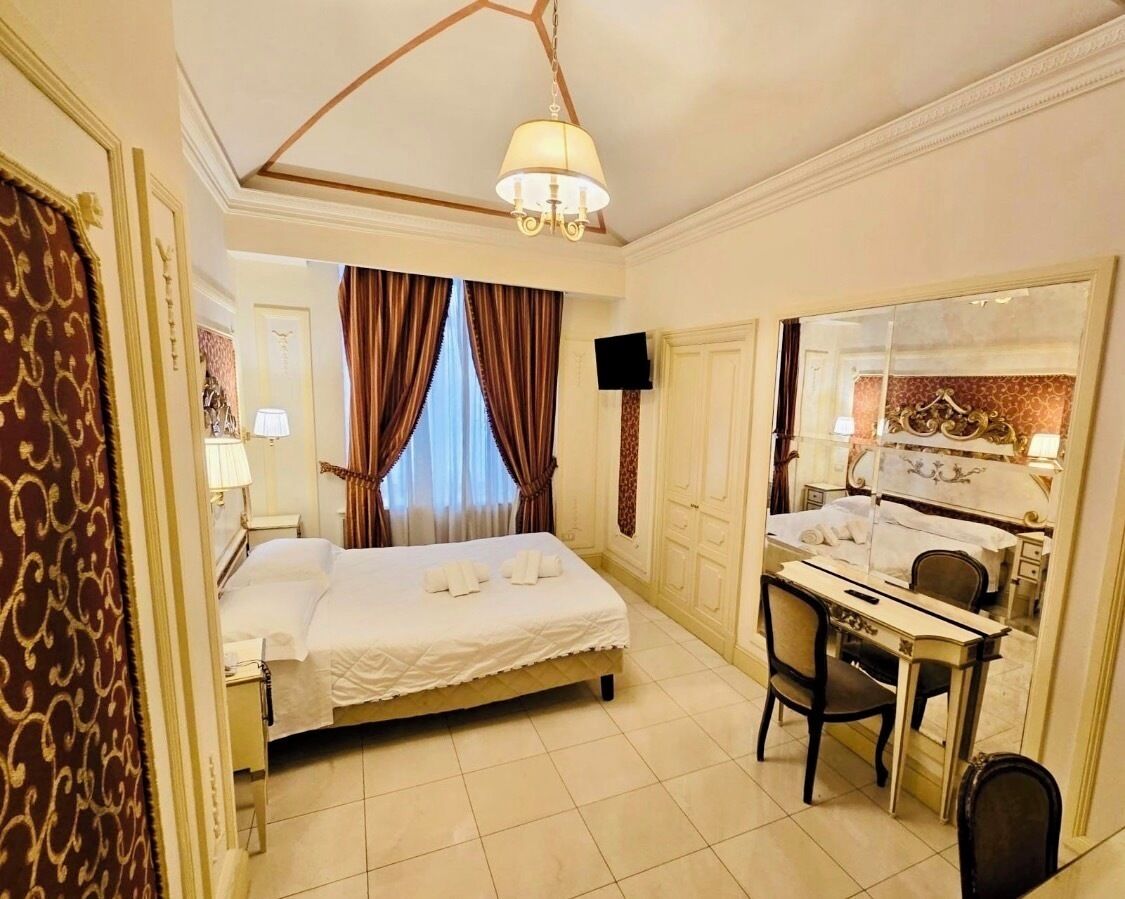 Une chambre d’hôtel comprenant un lit, un bureau, une chaise, un miroir, un lustre, des rideaux et un téléviseur.