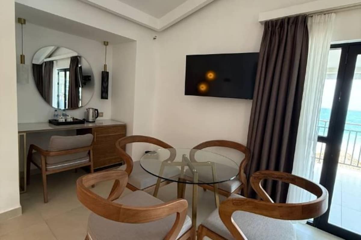 Habitación de hotel con zona de comedor, televisor montado en la pared y vistas al exterior.