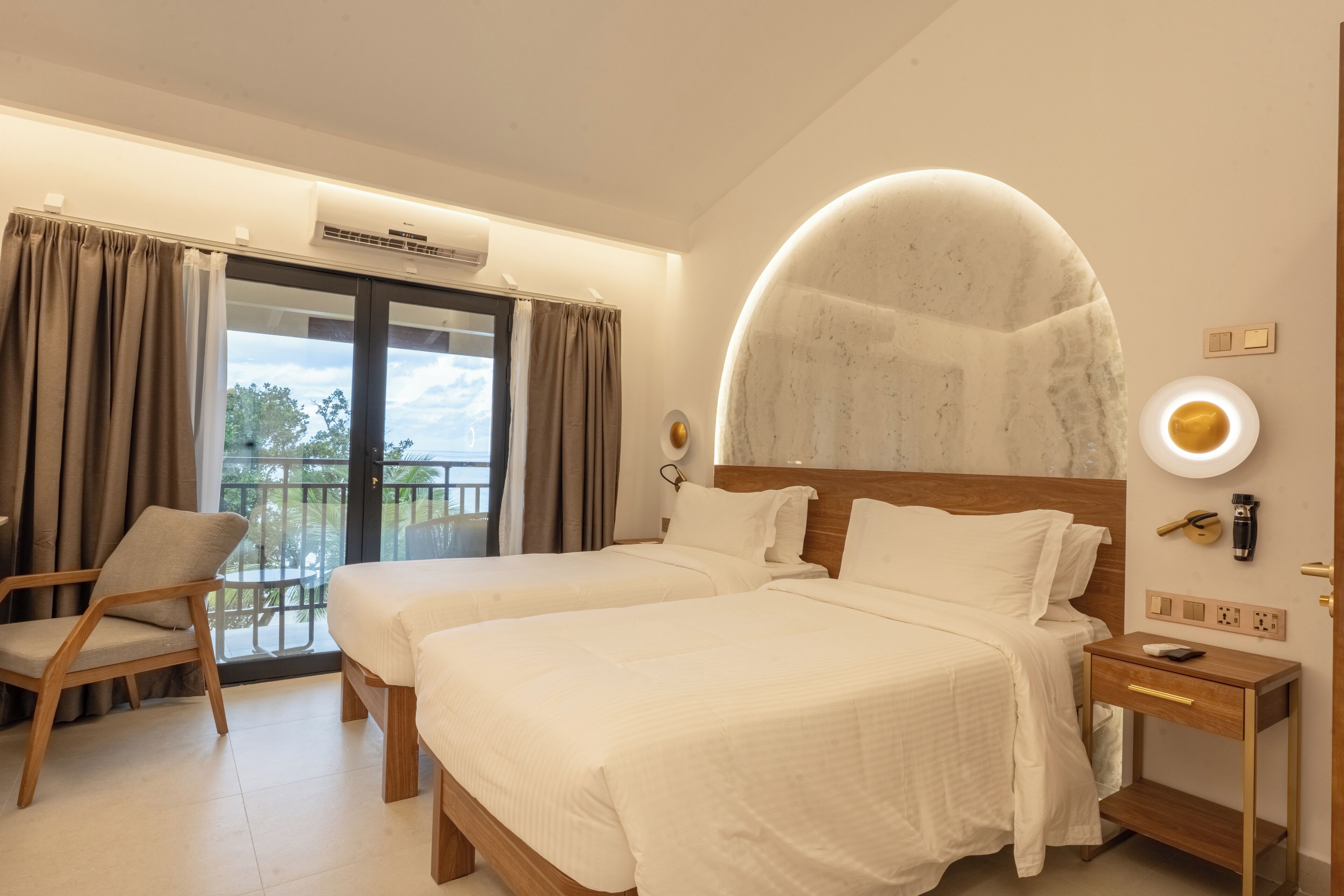 Habitación de hotel con dos camas, una silla, una mesita de noche, una lámpara de pared y un ventanal con cortinas.