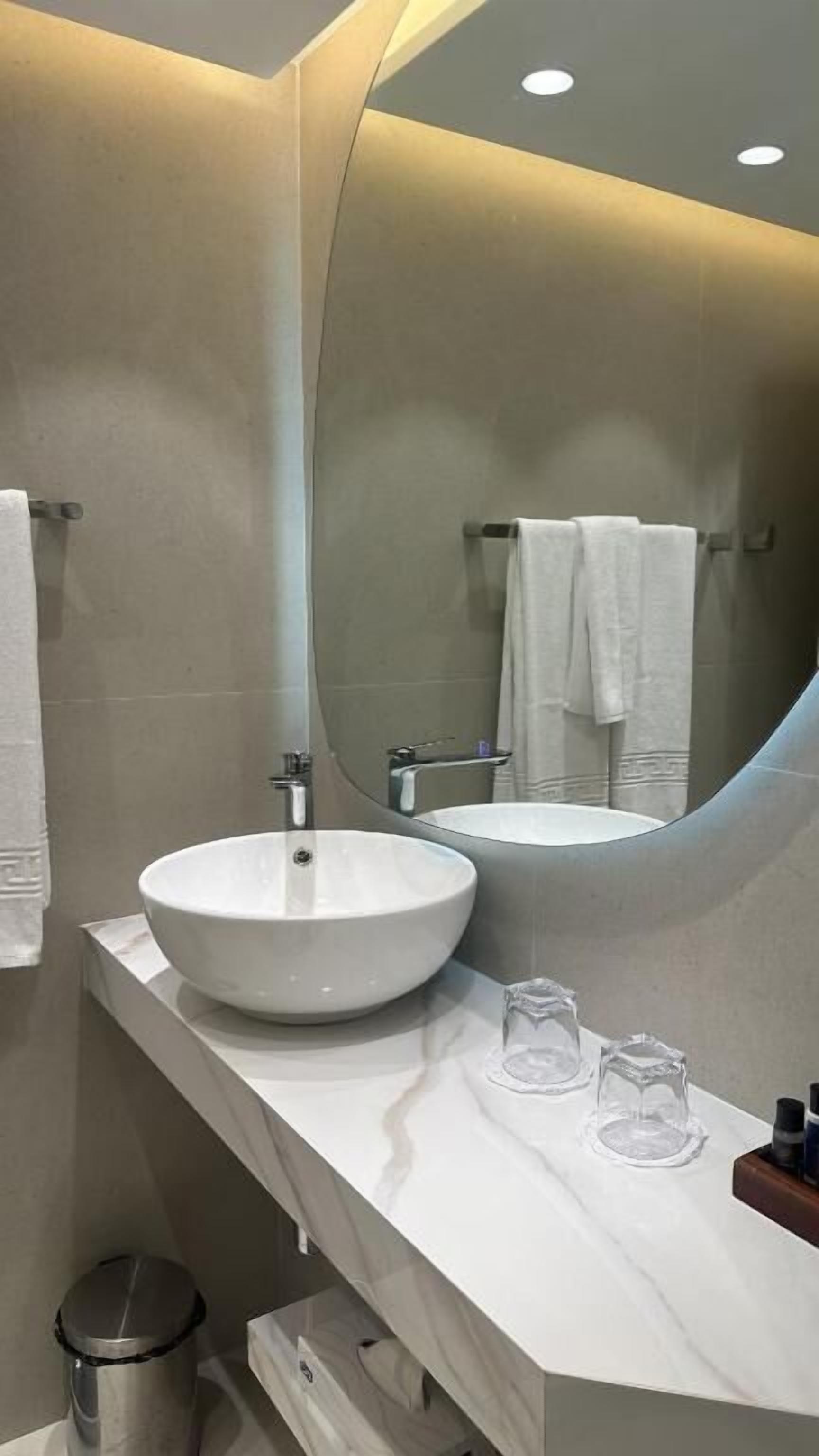 A modern bathroom with a large mirror, a white sink, and a vanity with a trash can.