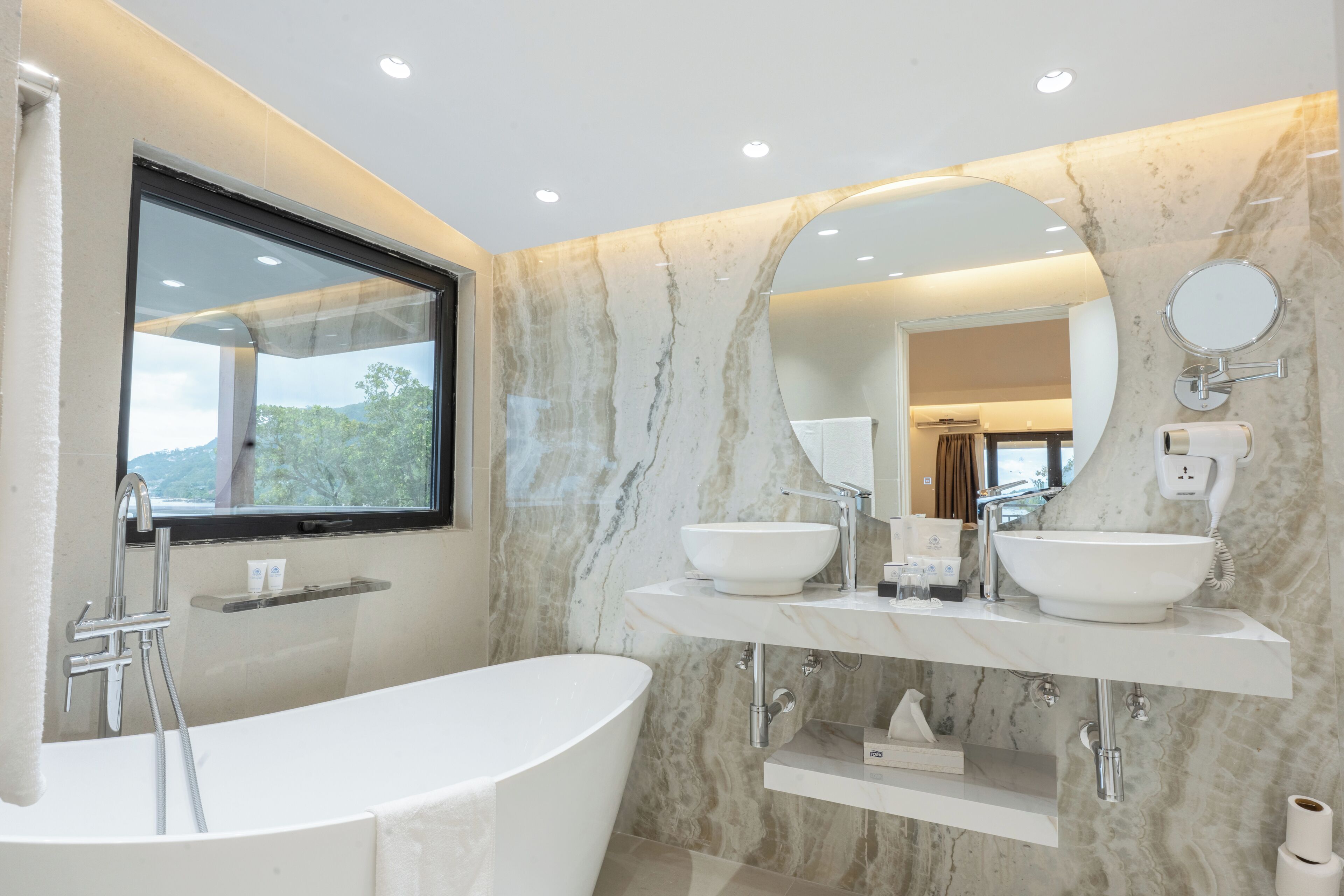 A modern bathroom with a freestanding bathtub, two vessel sinks, a large mirror, and a window with a view of greenery.