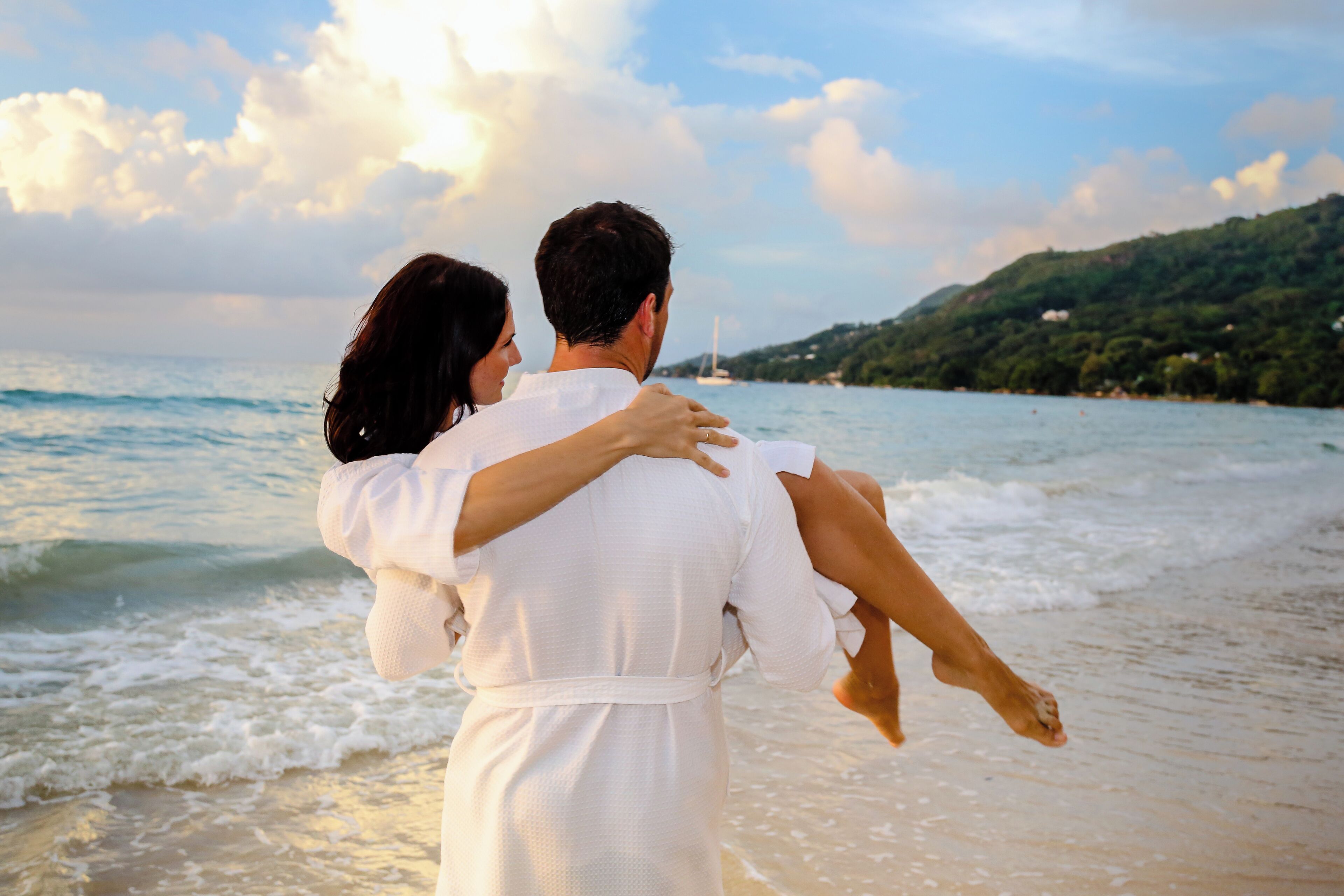 A couple embracing on a beach with waves and a hilly landscape in the background.