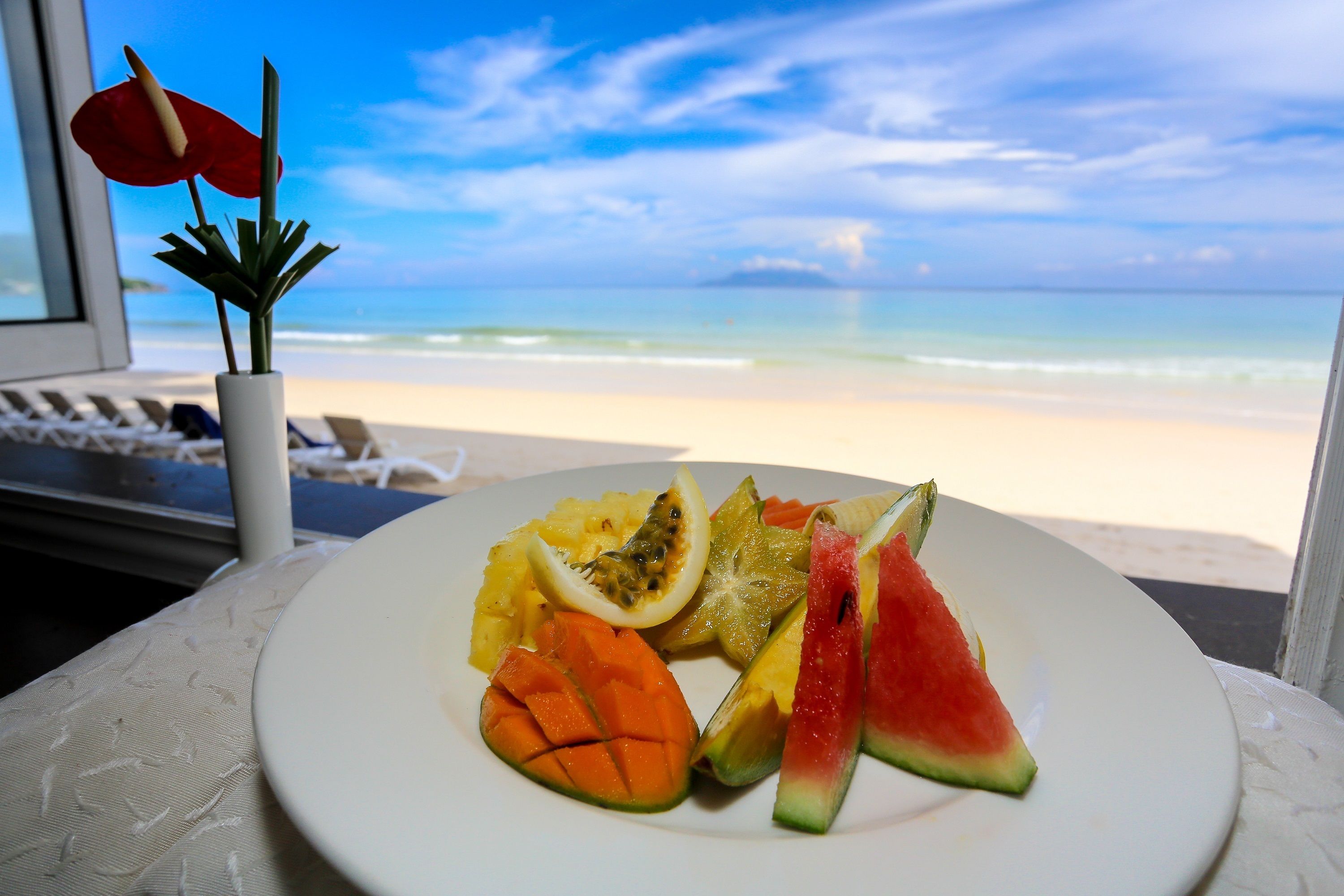 A plate of fruit on a table with a view of a beach and ocean through a window.