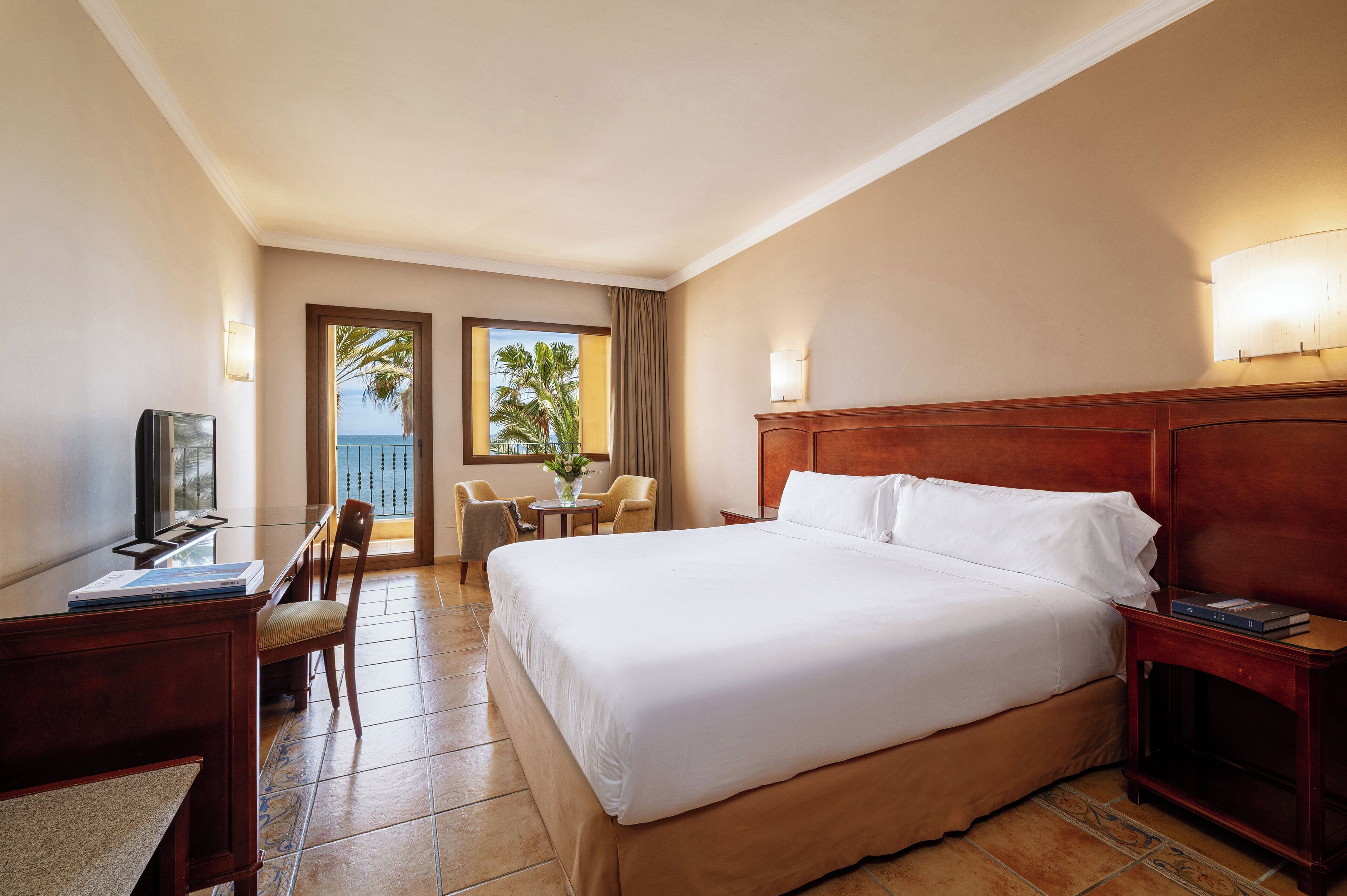 A hotel room with a large bed, a desk with a chair, a small table with a vase, and a view of palm trees through the window.