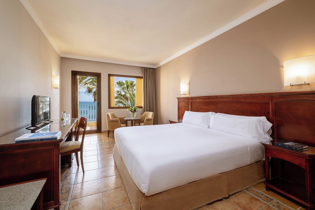 Hotelkamer met een groot bed, een bureau met een computer, een stoel, een kleine tafel met een vaas en uitzicht op het omliggende landschap.