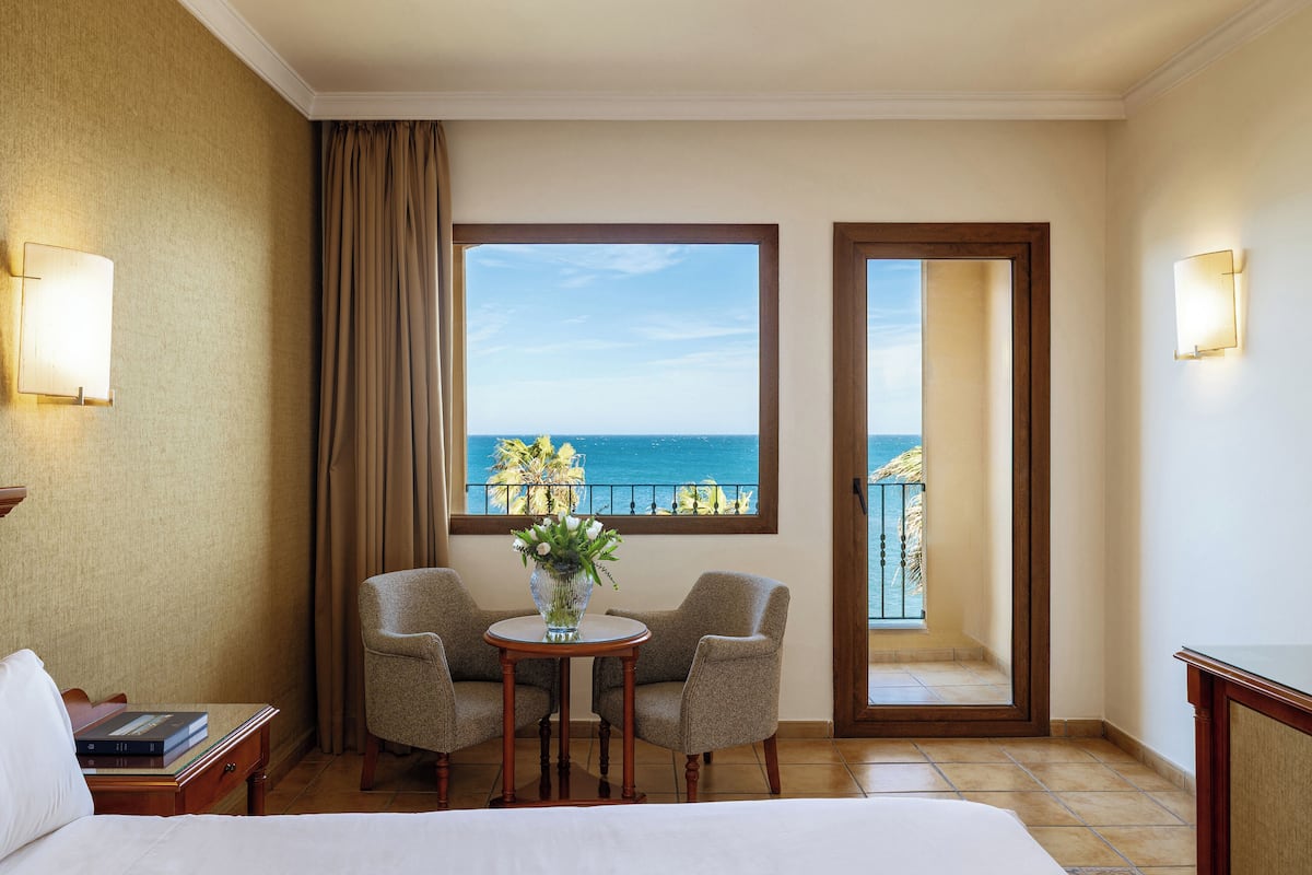 Hotelkamer met een bed, twee fauteuils, een bijzettafeltje met een vaas, een nachtkastje met boeken, een raam met uitzicht op zee, en een deur naar een balkon.