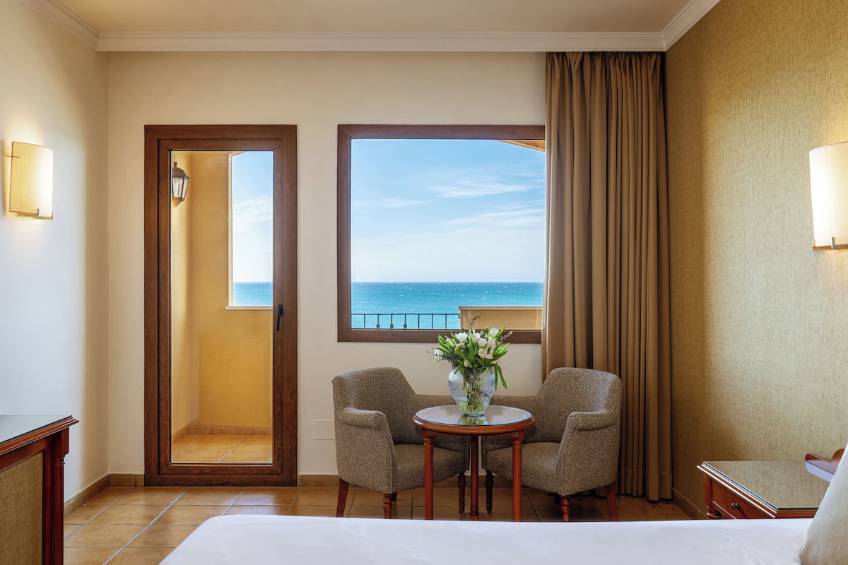 Een hotelkamer met een bed, twee fauteuils, een tafeltje met een vaas, een raam met uitzicht op zee, en een deur naar een buitenruimte.