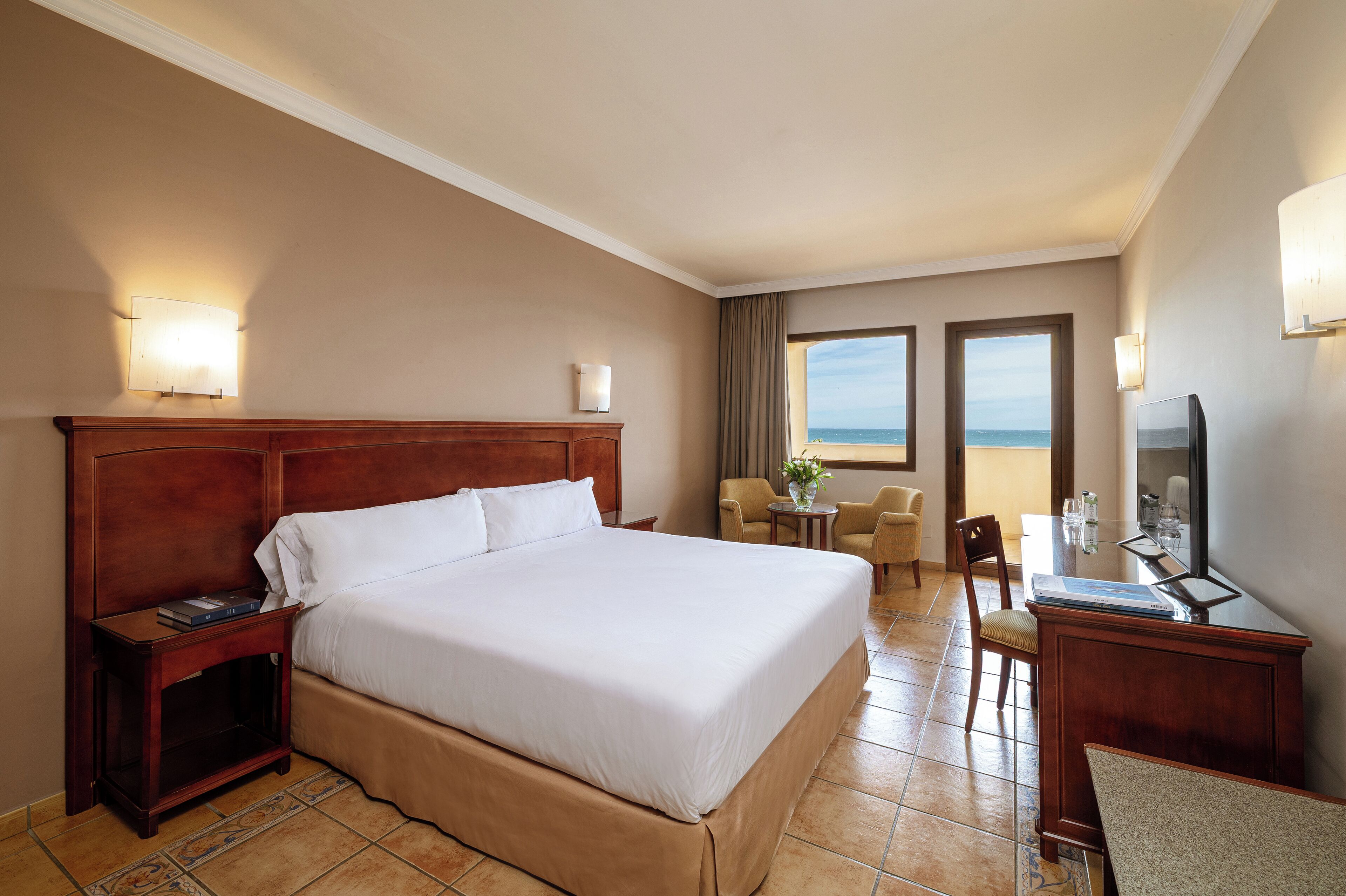 A hotel room with a large bed, a desk, a chair, a television, and a view of the ocean.
