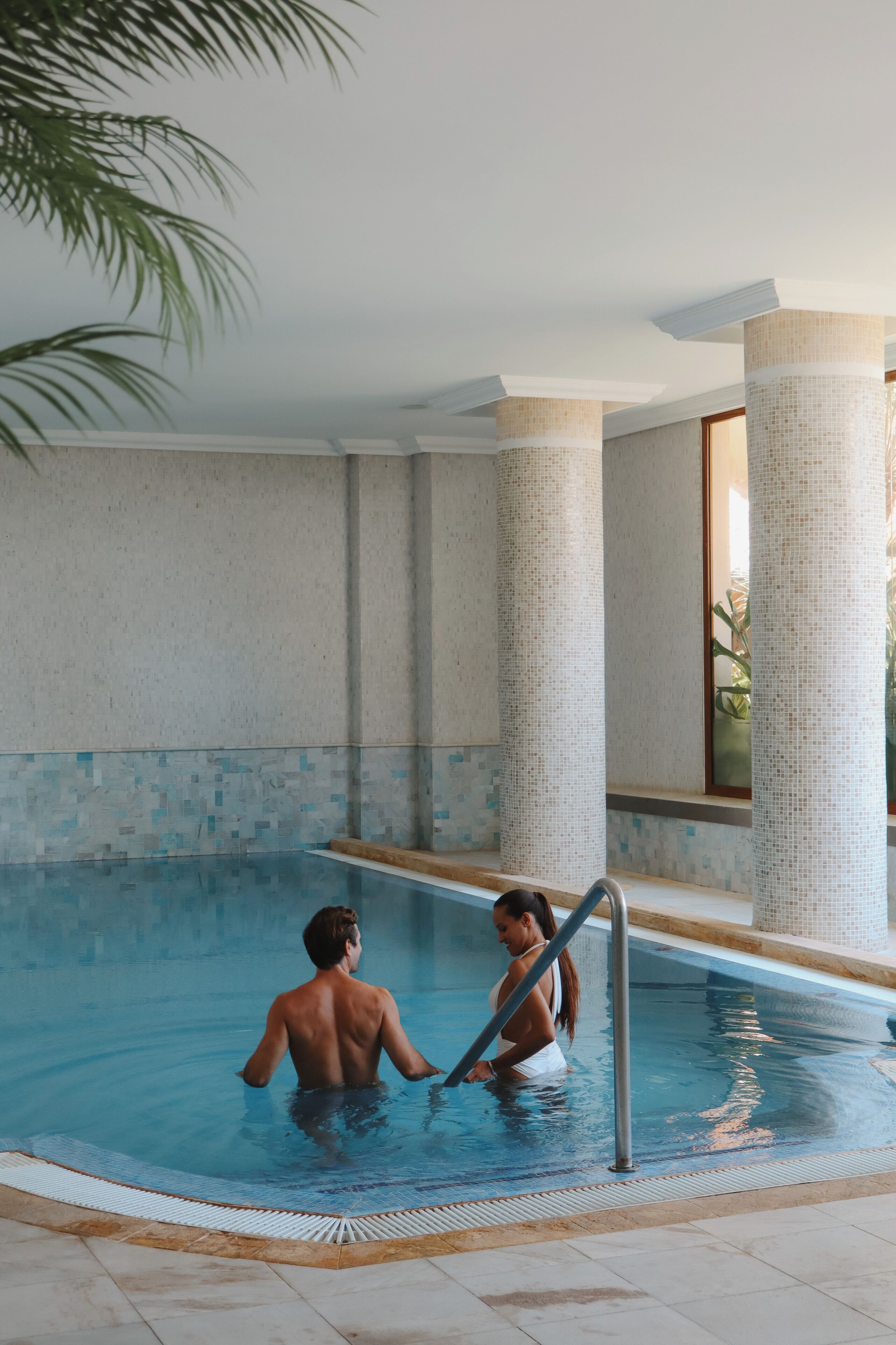 Una piscina interior con paredes de azulejos y columnas, borde de piscina transparente y una palmera al fondo.