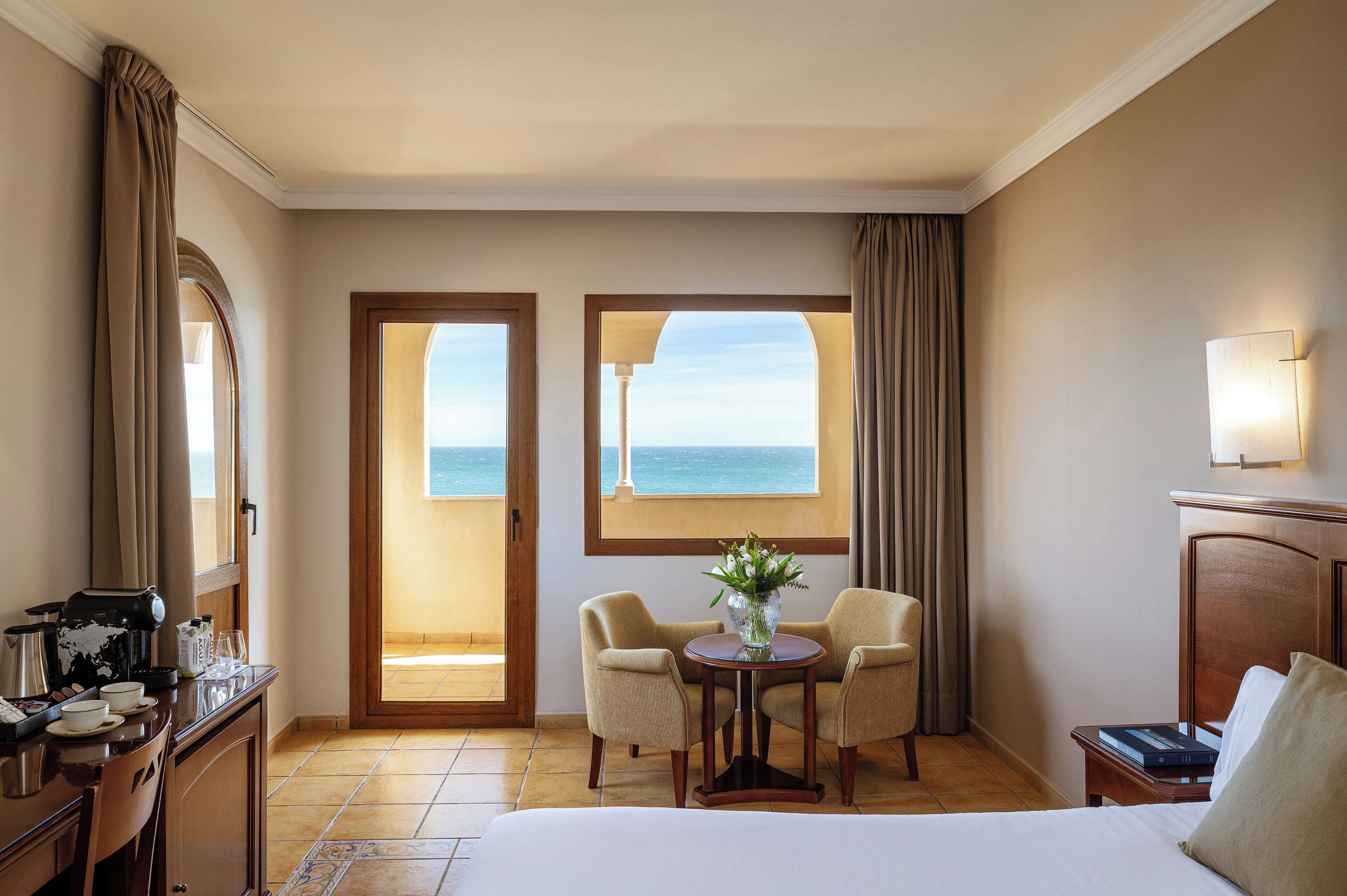 Une chambre d’hôtel avec un lit, un bureau doté d’une machine à café, deux chaises, une table avec un vase, et une vue sur la mer par la fenêtre.