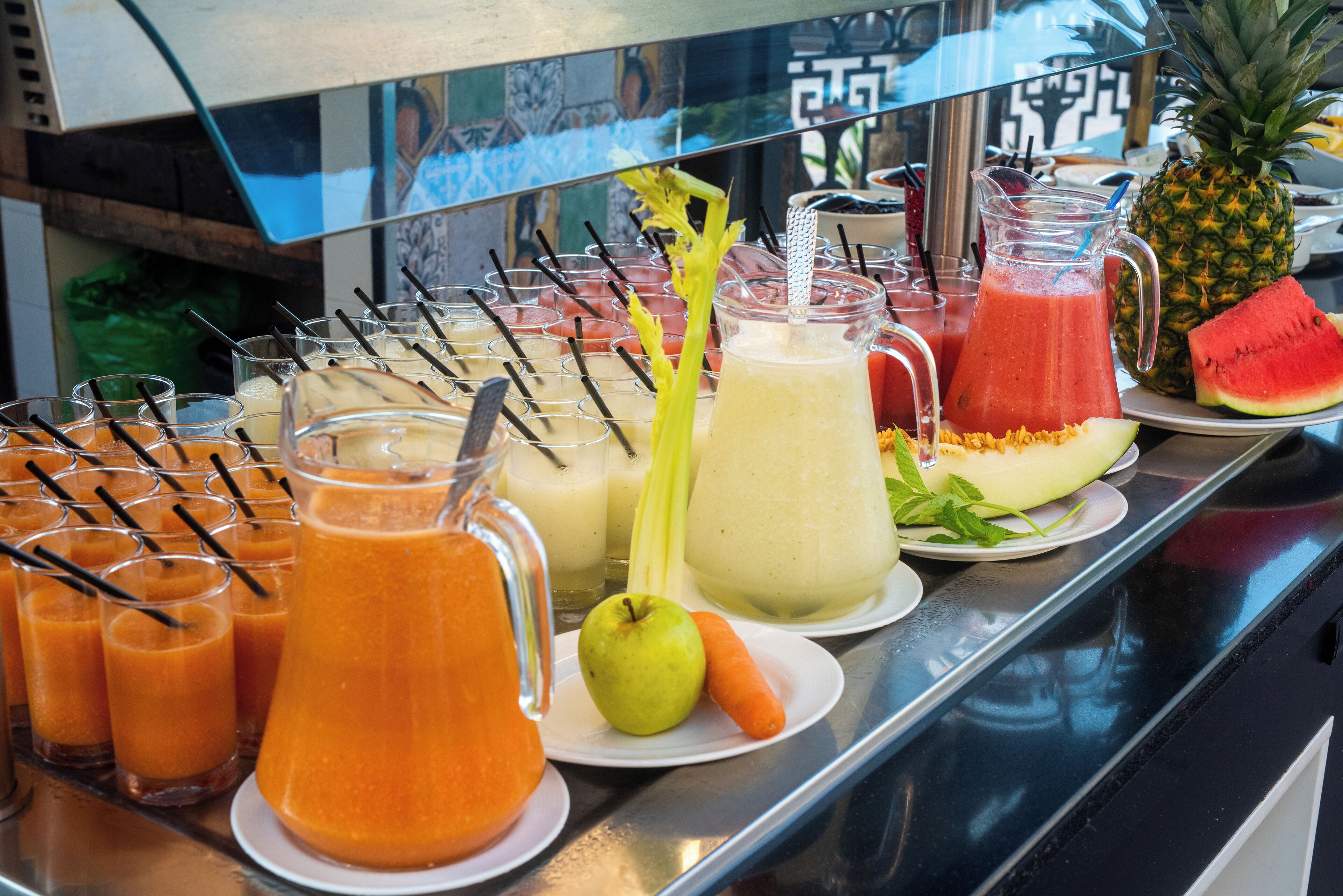 A buffet with various fruit juices, a pitcher of watermelon juice, a pineapple, and a melon on a counter.
