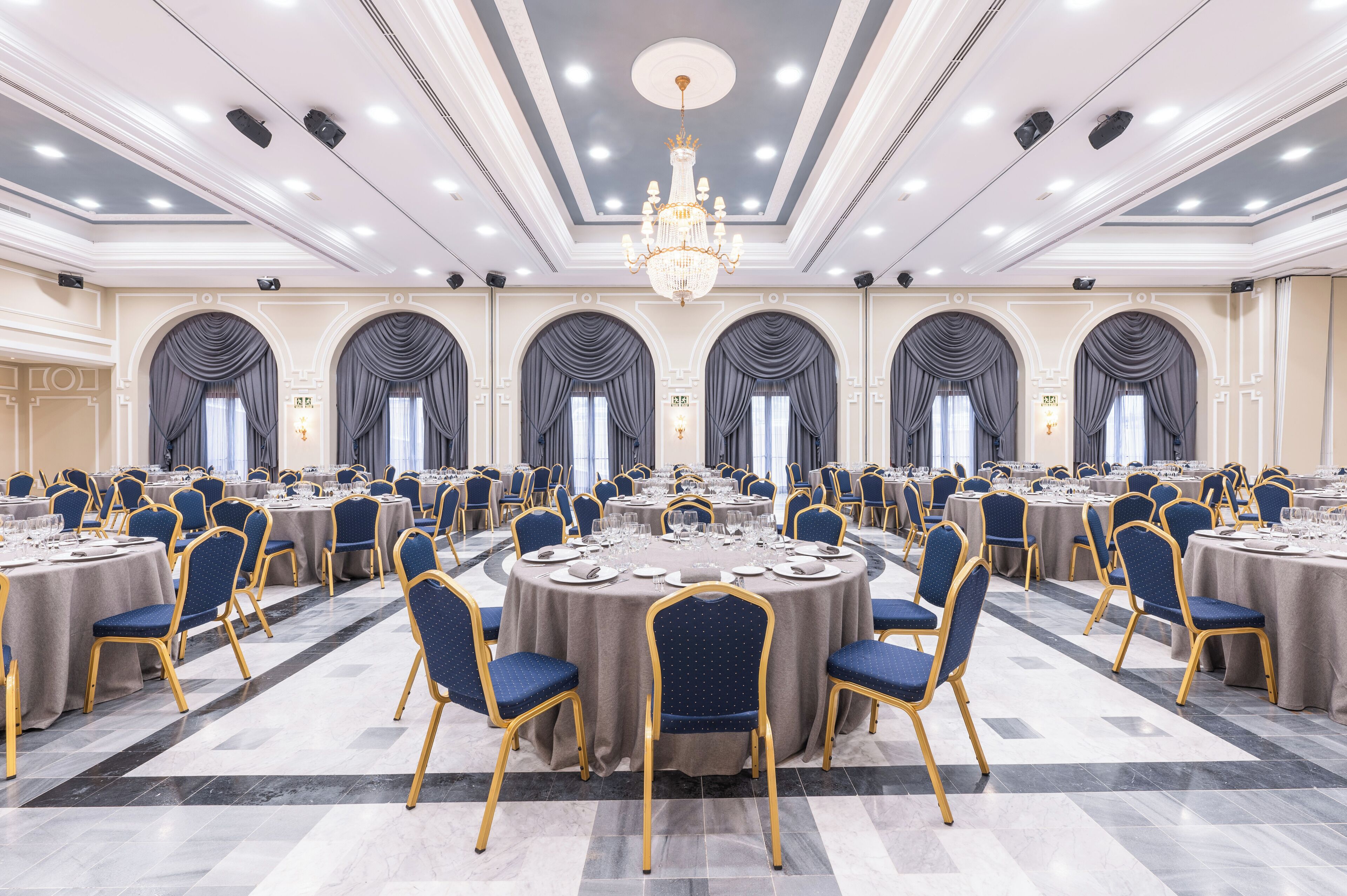 A banquet hall with round tables, blue chairs, and chandeliers.