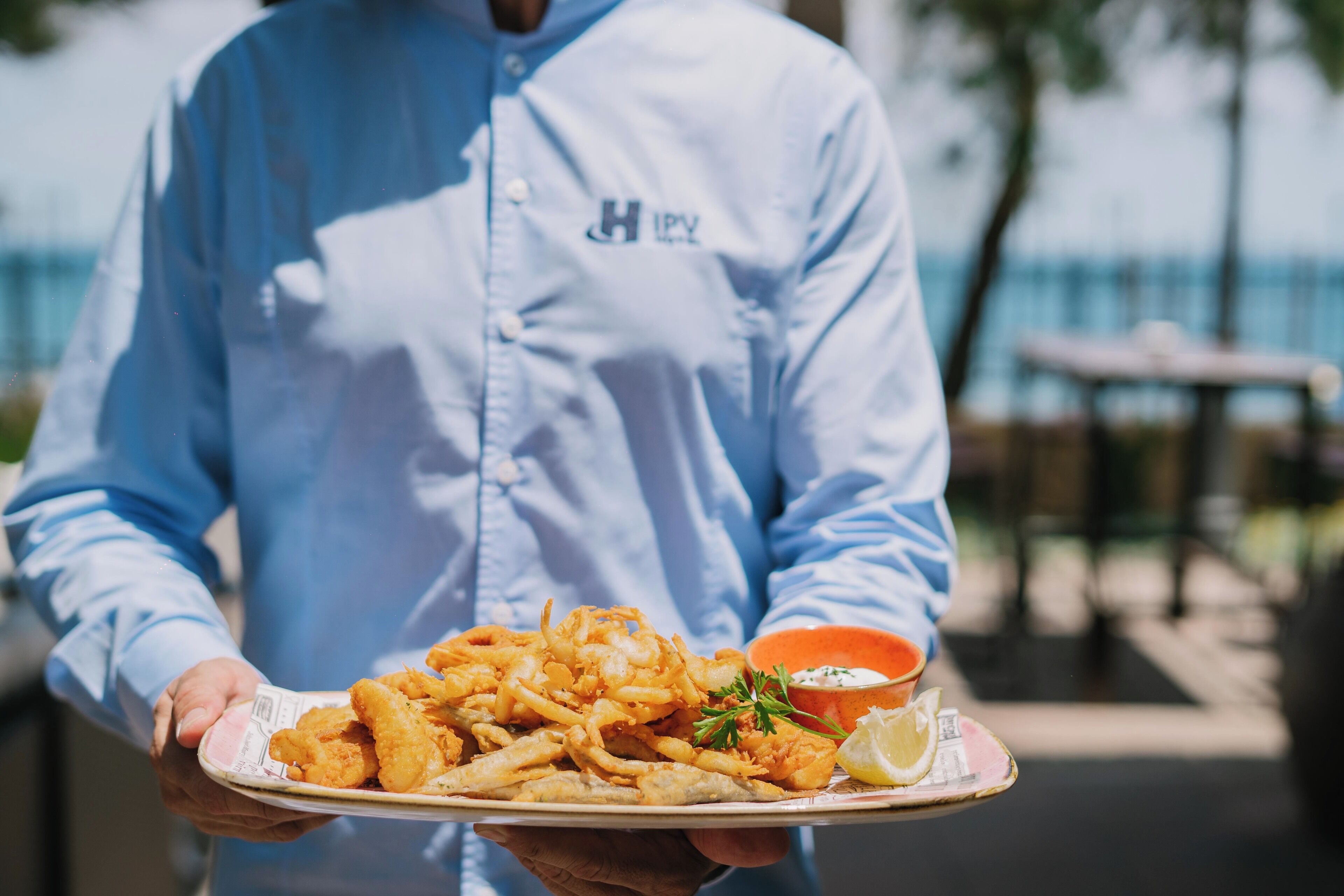 Une personne vêtue d’un uniforme bleu, tenant une assiette de nourriture frite et un petit récipient de sauce.