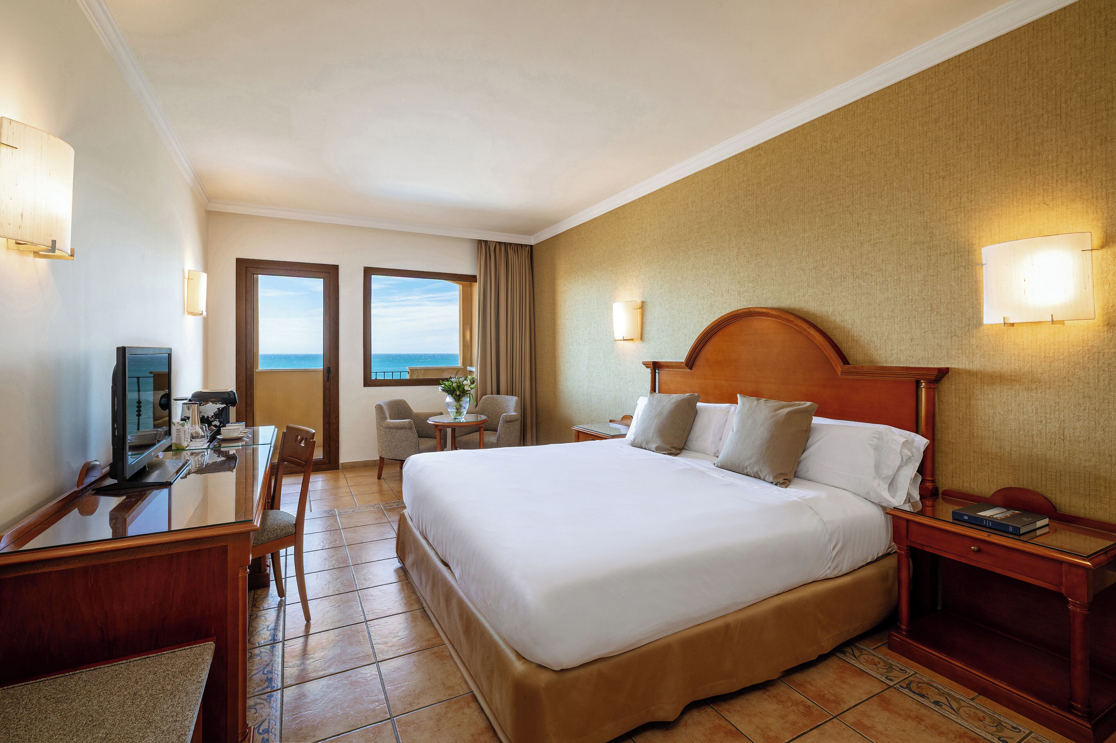Une chambre d’hôtel avec un grand lit, un bureau avec un ordinateur, une chaise, une petite table avec une plante et une vue sur l’océan par la fenêtre.