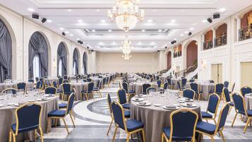 Une salle de banquet avec des tables rondes, des chaises recouvertes d’un tissu bleu, des lustres et un grand escalier.