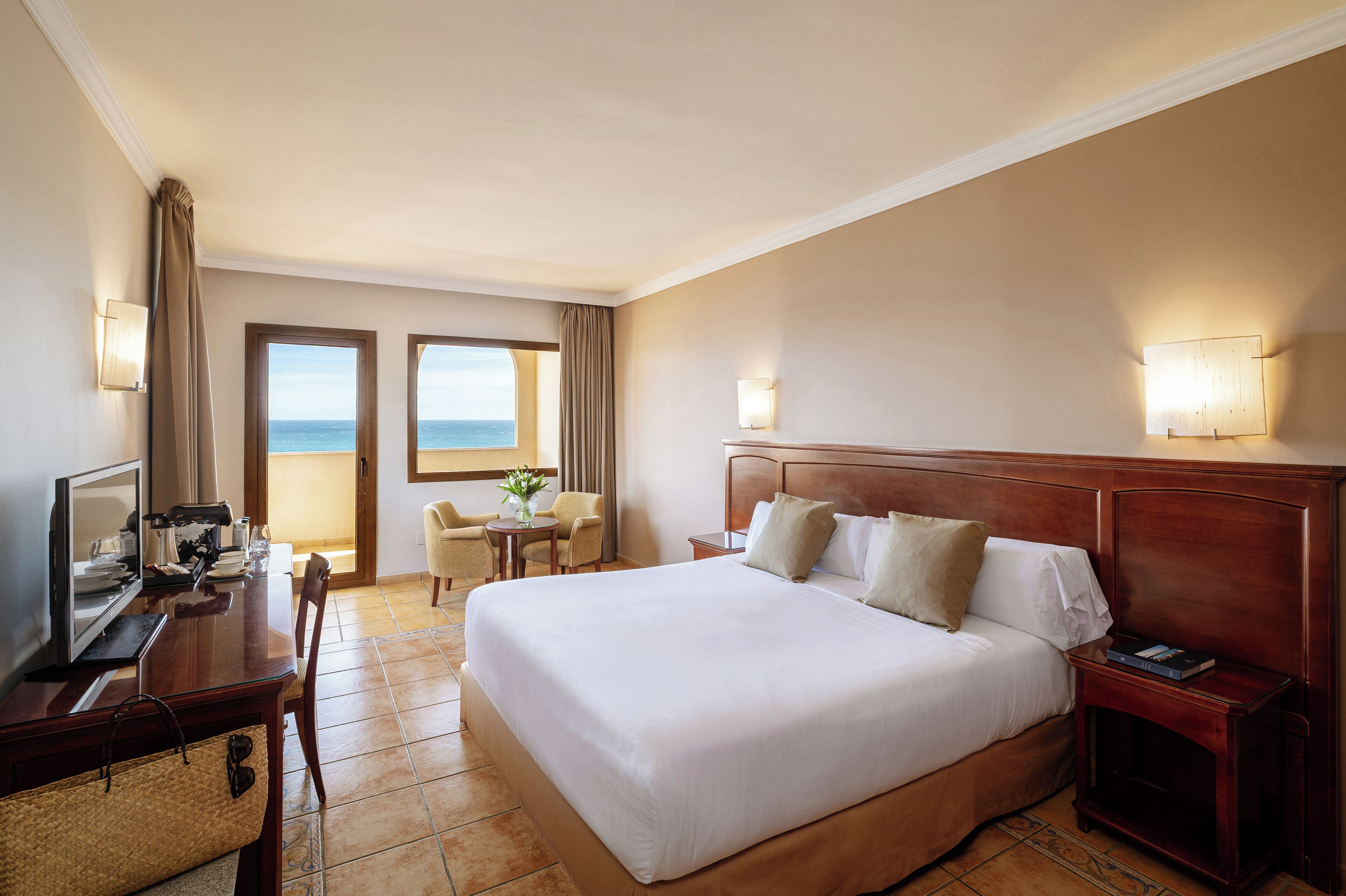 Habitación de hotel con una cama grande, un escritorio con una computadora, una silla, una mesita con un florero y vista al mar a través de una ventana.
