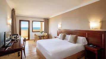 Habitación de hotel con una cama grande, un escritorio con una computadora, una silla, una mesita con un florero y vista al mar a través de una ventana.