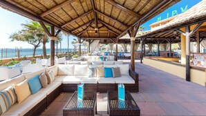 Un espacio de estar techado al aire libre con mobiliario de mimbre, vistas al mar y un edificio con la señalización "HIPV".