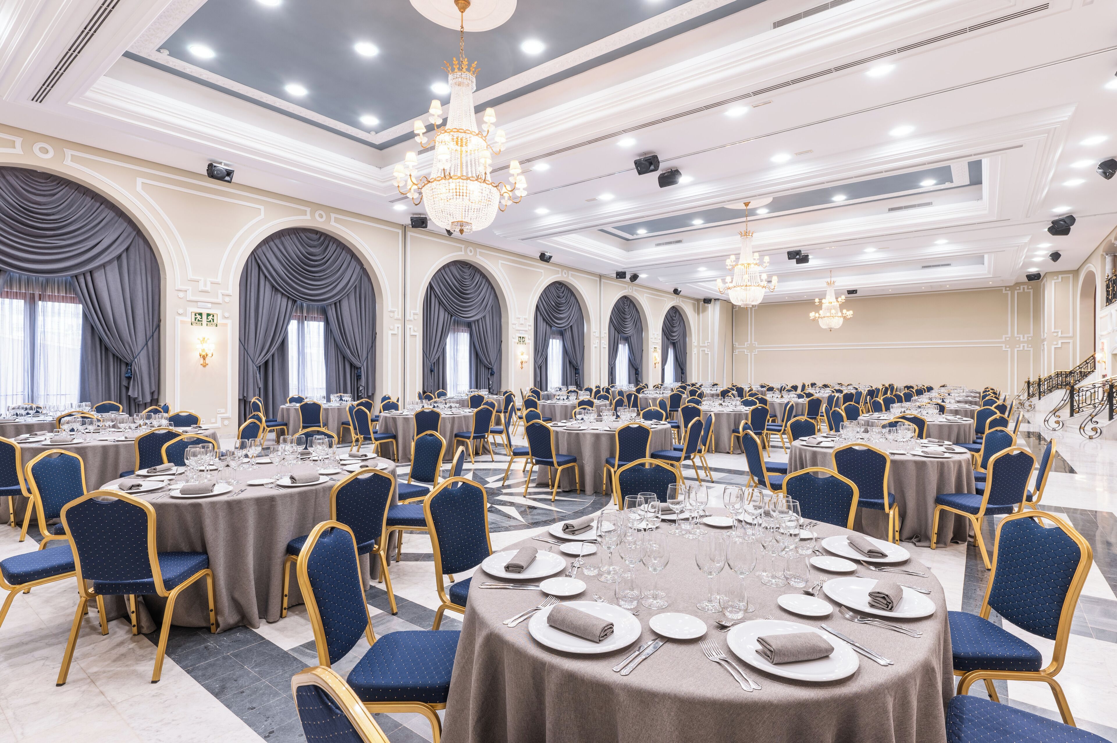 A banquet hall with round tables, blue upholstered chairs, chandeliers, and a grand staircase.