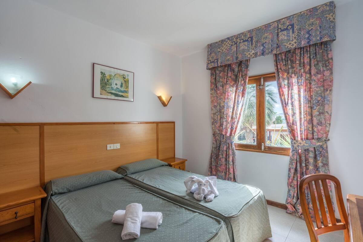 Une chambre à coucher avec une tête de lit en bois, deux lits, une table de chevet, une lampe fixée au mur, une fenêtre avec des rideaux à motifs floraux et un tableau encadré accroché au mur.