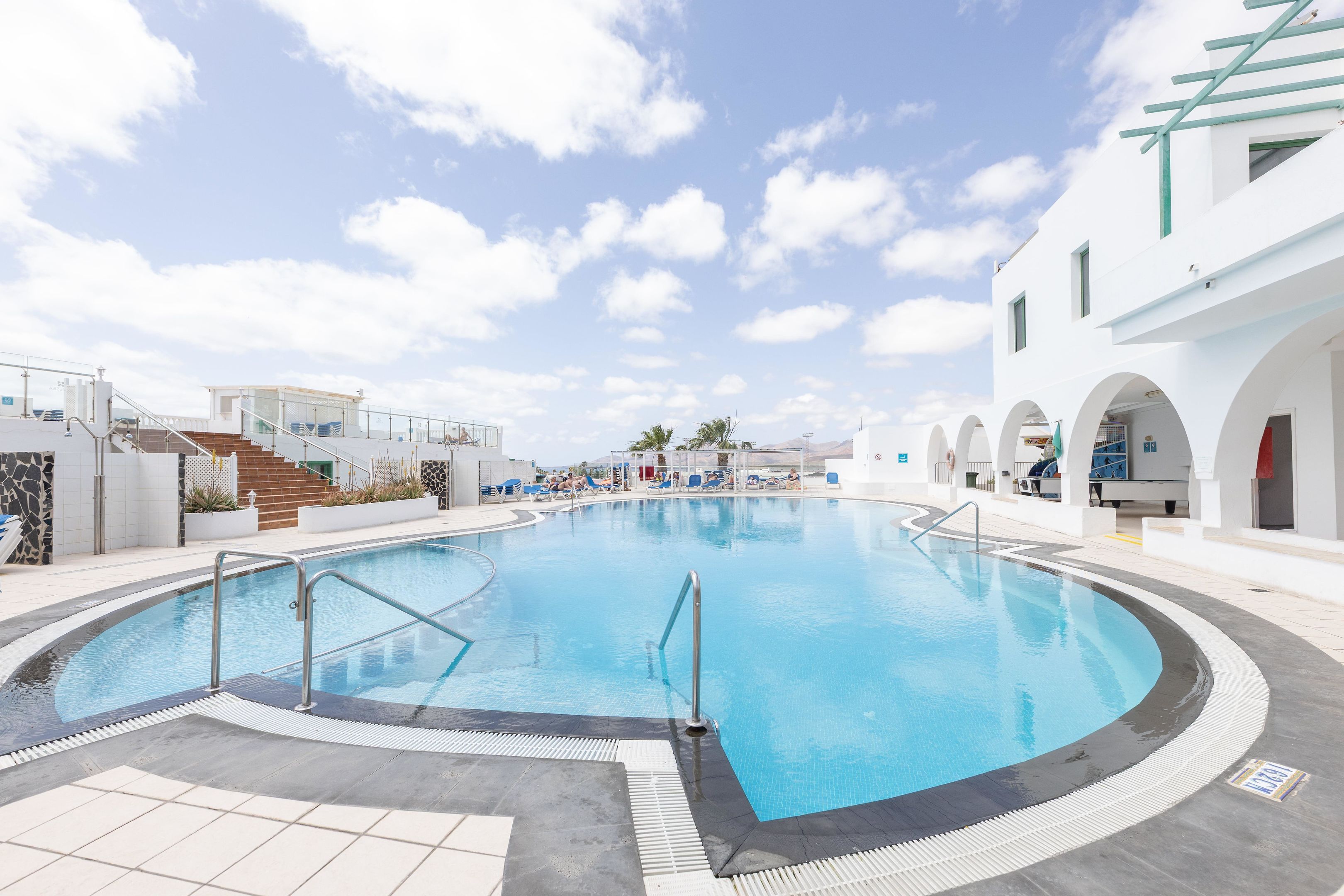 A swimming pool with a clear blue water surface, surrounded by a tiled deck and a white building with arches.