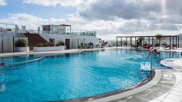 A large outdoor swimming pool with clear blue water, surrounded by lounge chairs and a covered seating area. - BLUESEA Los Fiscos (Tías)