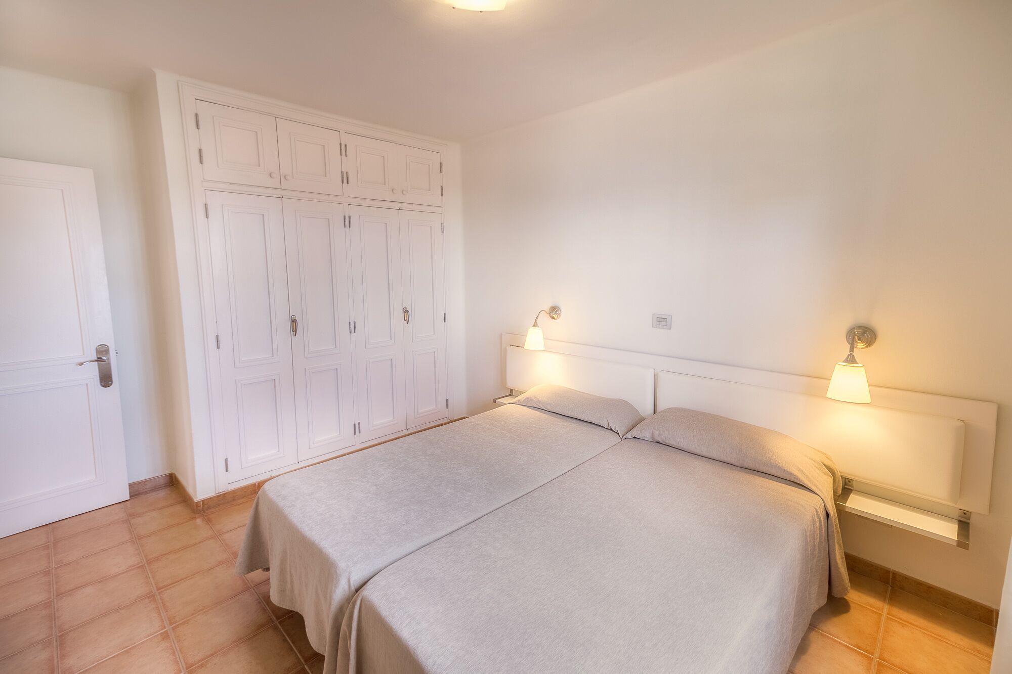 Une chambre à coucher avec un lit double, deux lampes de chevet, une armoire à plusieurs portes et un sol carrelé.