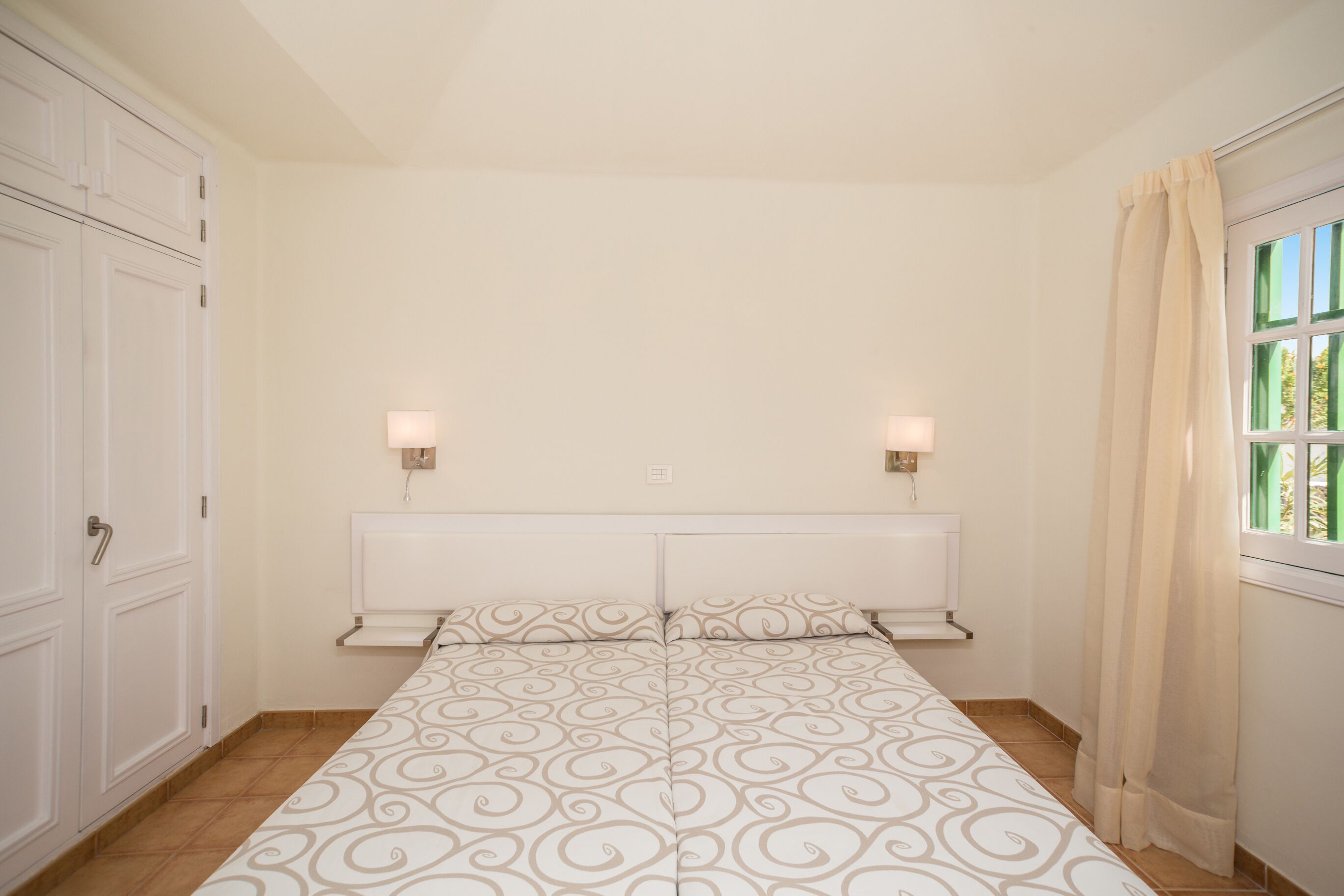 A bedroom with a bed, two bedside lamps, a window with curtains, and a wardrobe.