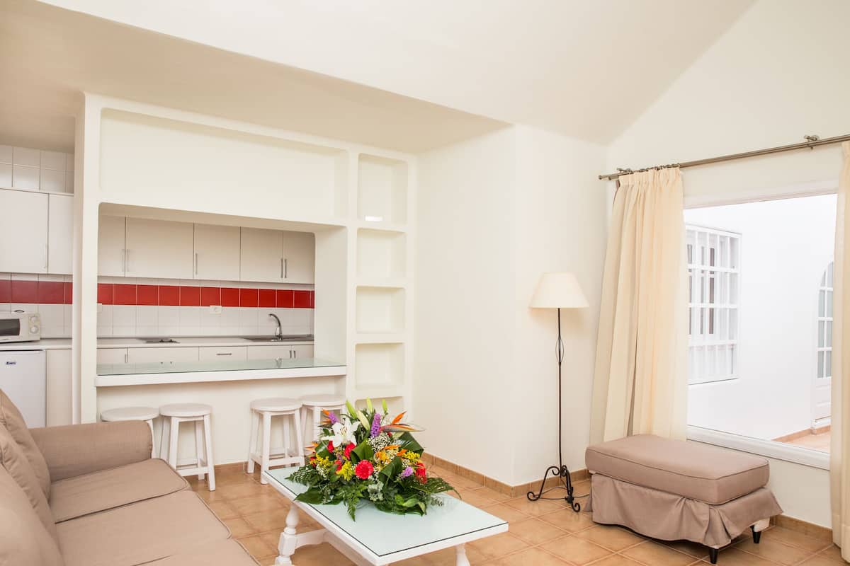 Een moderne woonkamer met een bank, een eettafel met een bloemstuk, een keuken met witte kastjes en een raam met gordijnen.