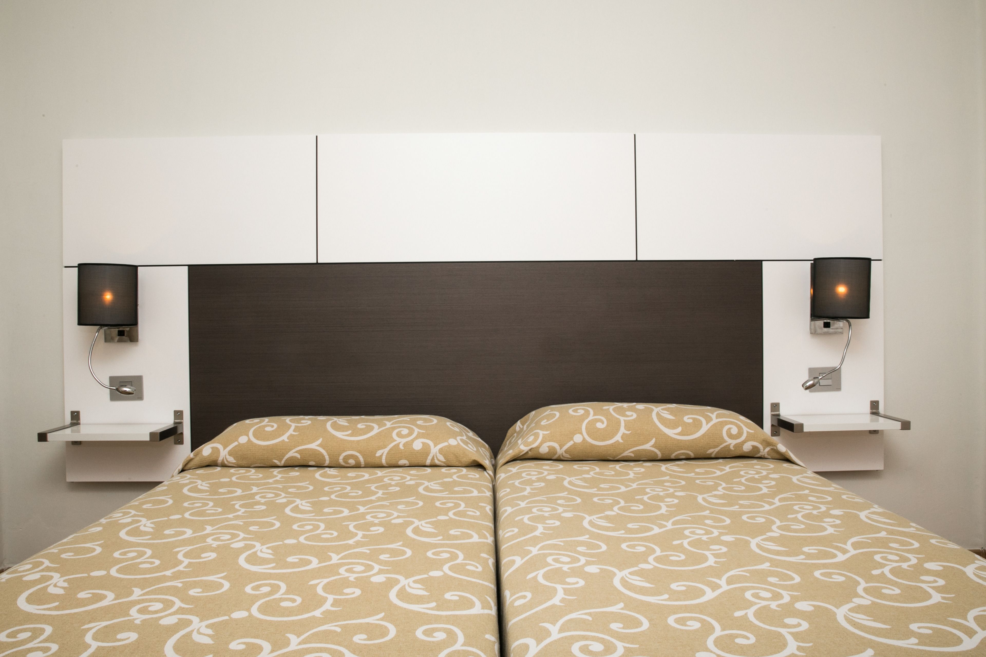 Un lit double avec une couvre-lit à motifs, une tête de lit sombre et deux tables de chevet fixées au mur, chacune équipée d’une lampe.