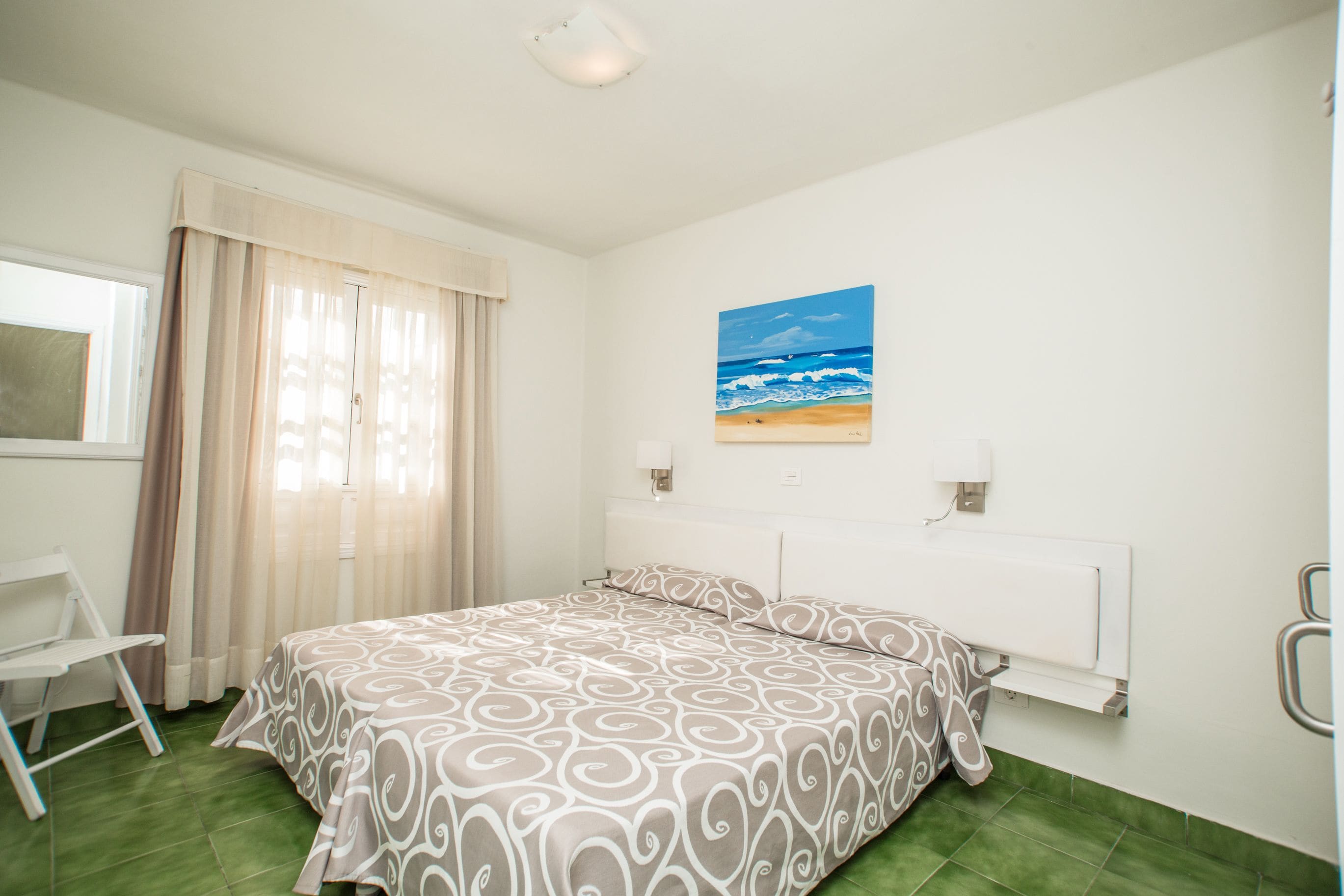 Un dormitorio con cama, una silla, un cuadro de una playa y una ventana con cortinas.