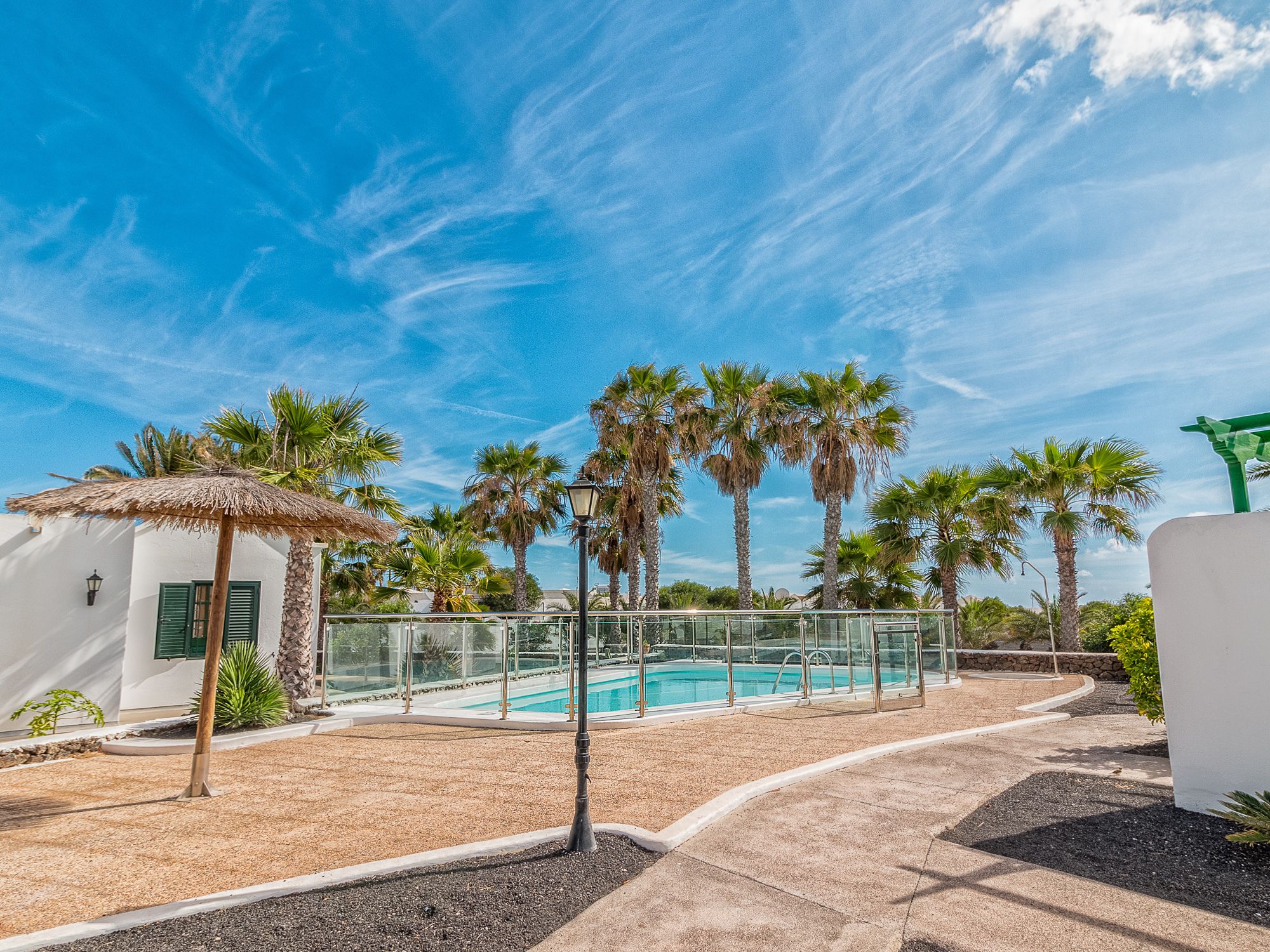 Zona de piscina amb palmeres, una construcció amb teulada de palla i un cel blau clar.