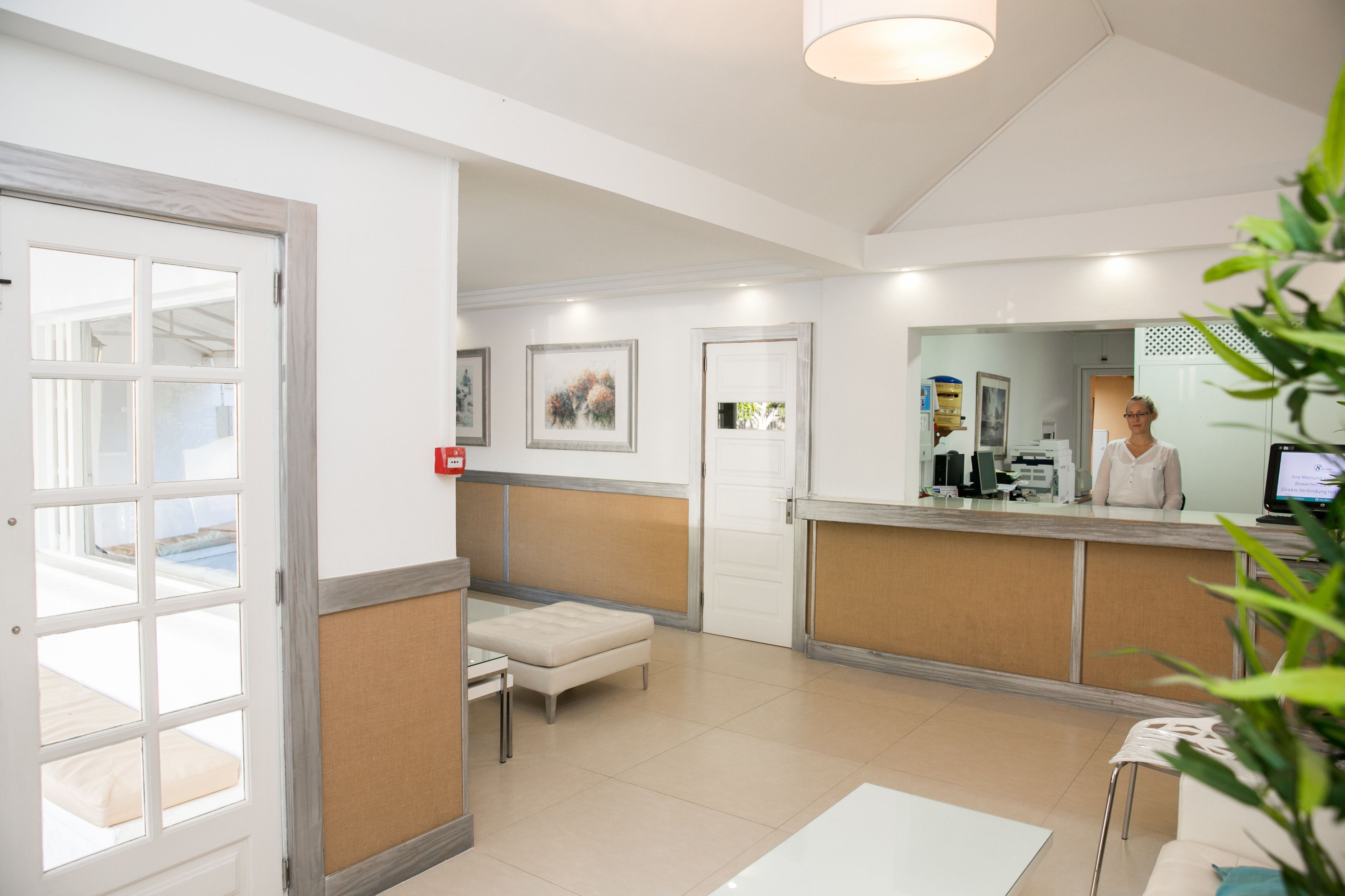 Un área de recepción con recepcionista, un mostrador, un espacio de espera con banca y una puerta que da a otra sala.