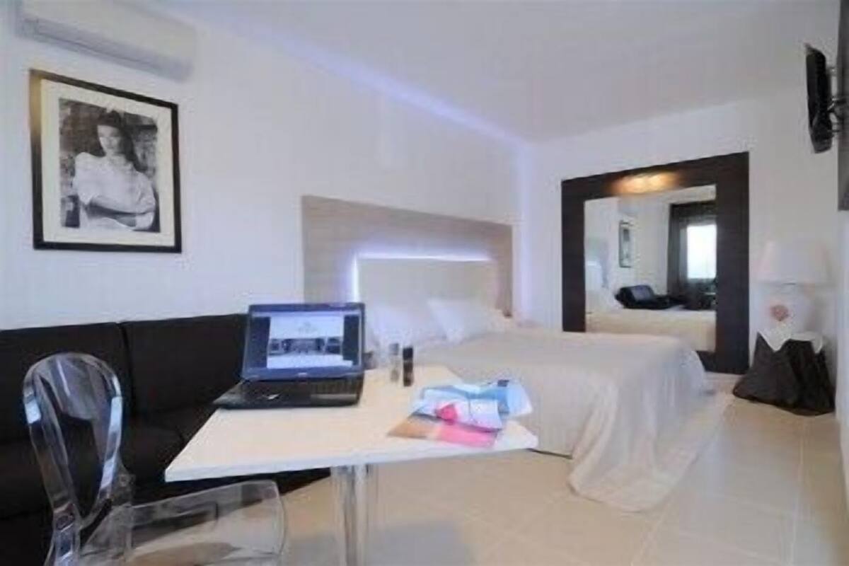 A hotel room with a bed, a desk with a laptop, a chair, and a bedside table with a lamp.