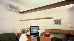 A small, well-lit room with a green sofa, a small table with a lamp, a bed with green bedding, a desk with a computer and a fruit basket, and a framed picture on the wall. - Hotel La Corte (Olbia)