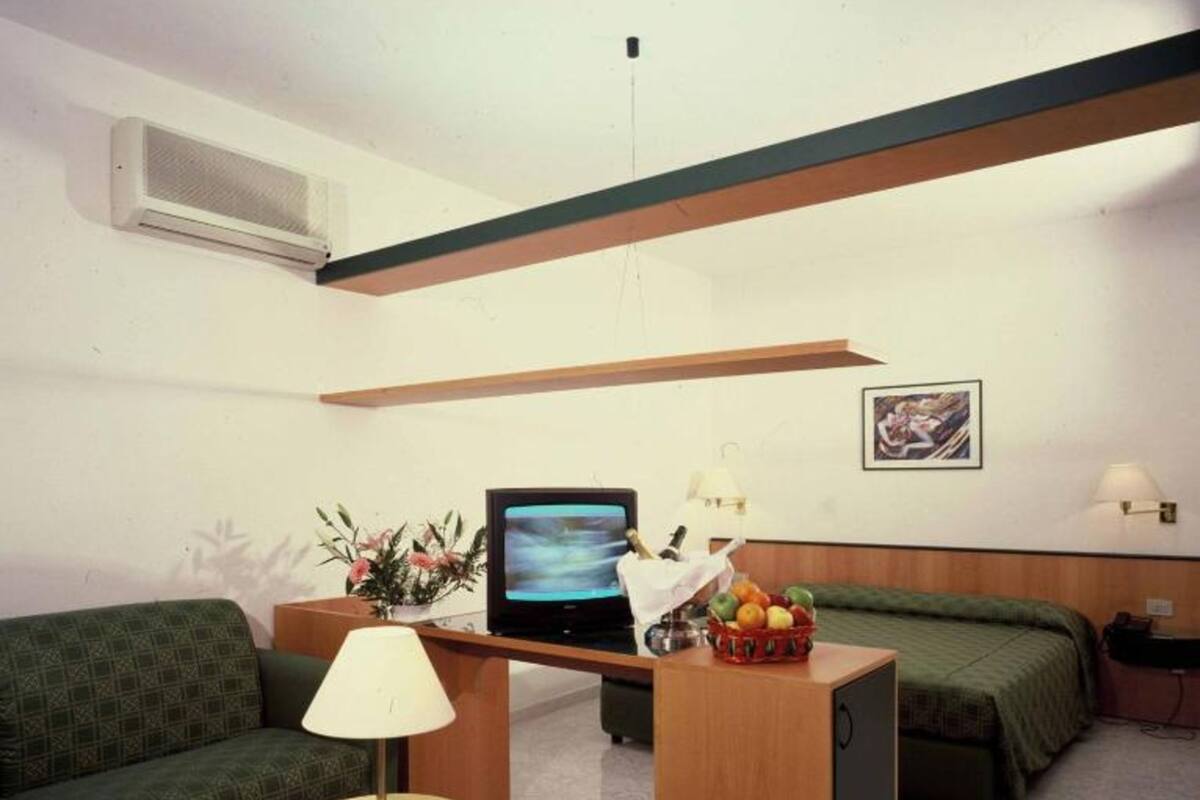 A small, well-lit room with a green sofa, a small table with a lamp, a bed with green bedding, a desk with a computer and a fruit basket, and a framed picture on the wall.
