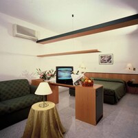 Una habitación pequeña y luminosa con un sofá verde, una mesita de noche con una lámpara, una cama con ropa de cama verde, un escritorio con una computadora y una cesta de frutas, y un cuadro en la pared.
