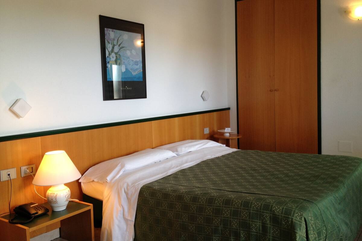 A hotel room with a bed, a nightstand with a lamp, a phone, a framed picture on the wall, and a wooden wardrobe.