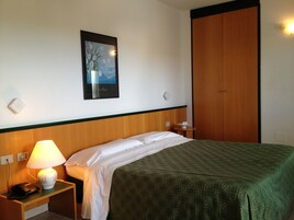 Una habitación de hotel con una cama, una mesita de noche con lámpara, un teléfono, un cuadro enmarcado en la pared y un armario de madera.