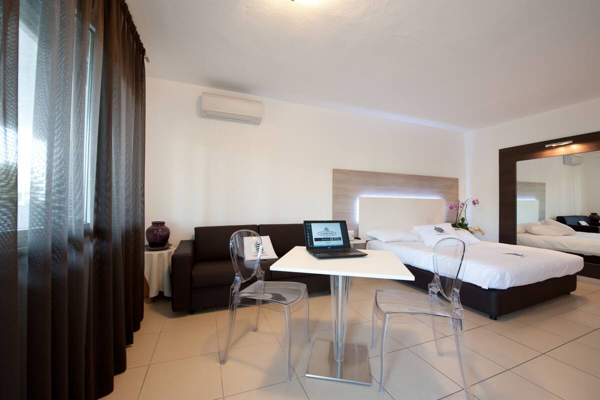 A hotel room with a bed, a desk with a laptop, a chair, a sofa, and a large mirror.