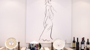 A minimalist art gallery with a large line drawing on the wall, a table with wine bottles, and decorative plates.