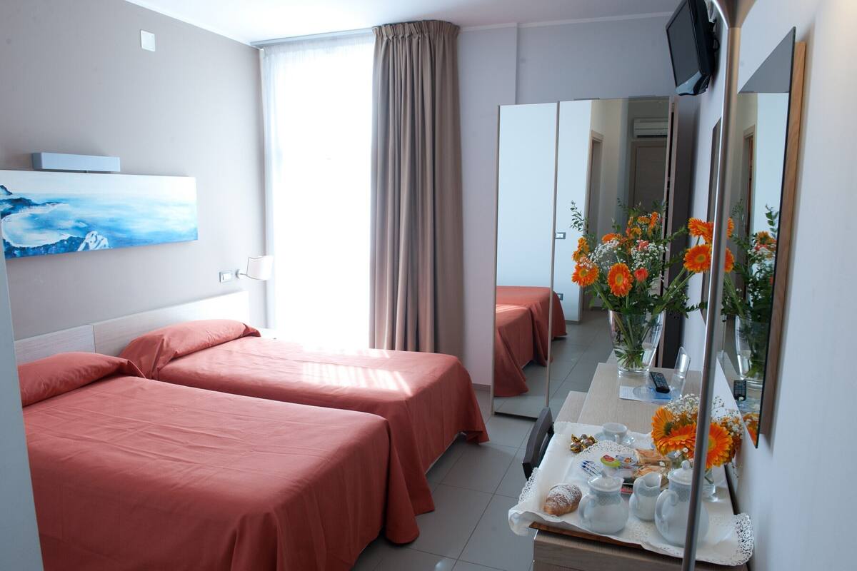 A hotel room with two beds, a large mirror, a painting on the wall, and a table with a vase of flowers.