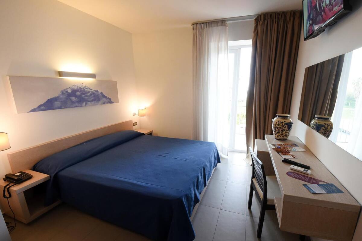 A hotel room with a bed, bedside table, lamp, artwork, and a desk with a vase and remote controls.