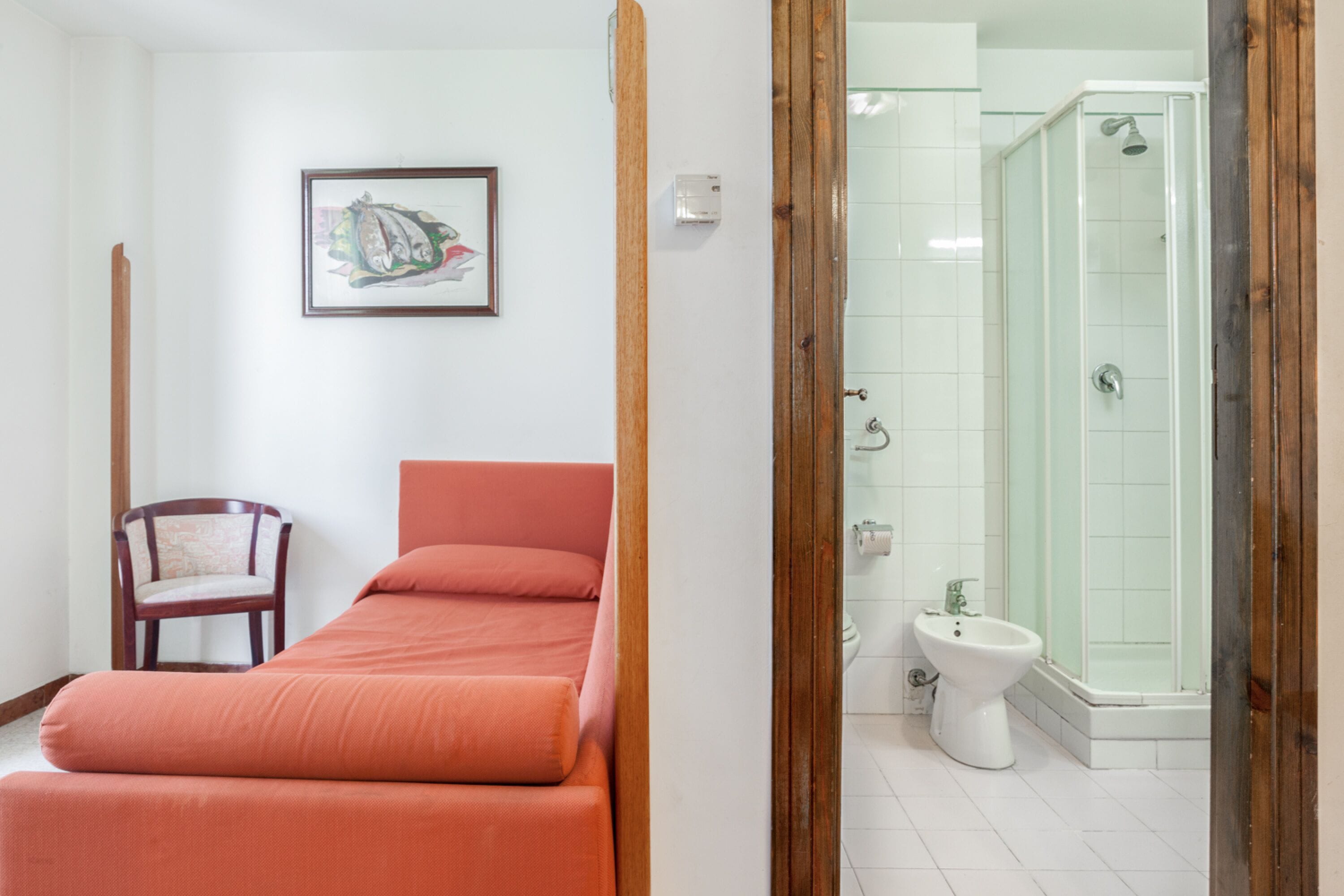 Una habitación con cama, una silla, un cuadro en la pared y un baño con ducha e inodoro.